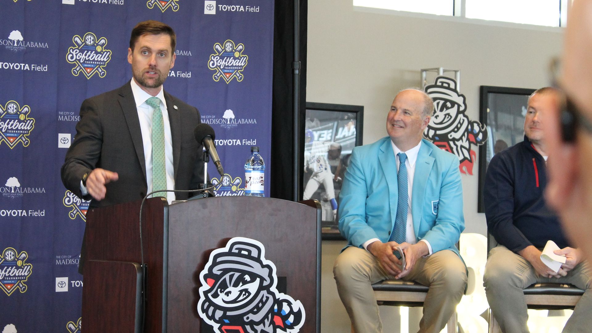 Man in a dark suit speaks at a podium during a softball event, blue backdrop covered with logos. A man in a light blue blazer sits to the right, smiling, with others nearby.