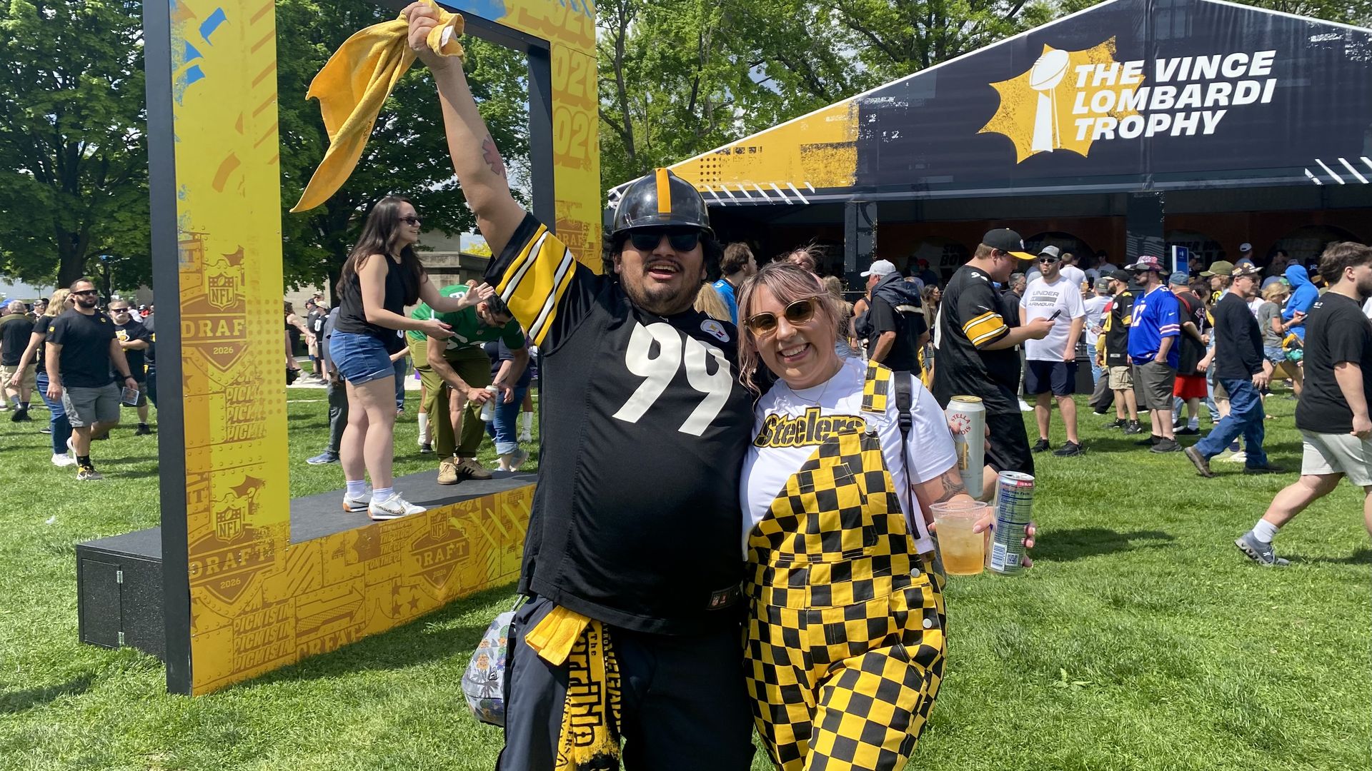 Two Steelers fans in black and yellow outfits pose with a raised scarf, smiling by a yellow NFL Draft frame and a large tent reading "The Vince Lombardi Trophy" at an outdoor event.