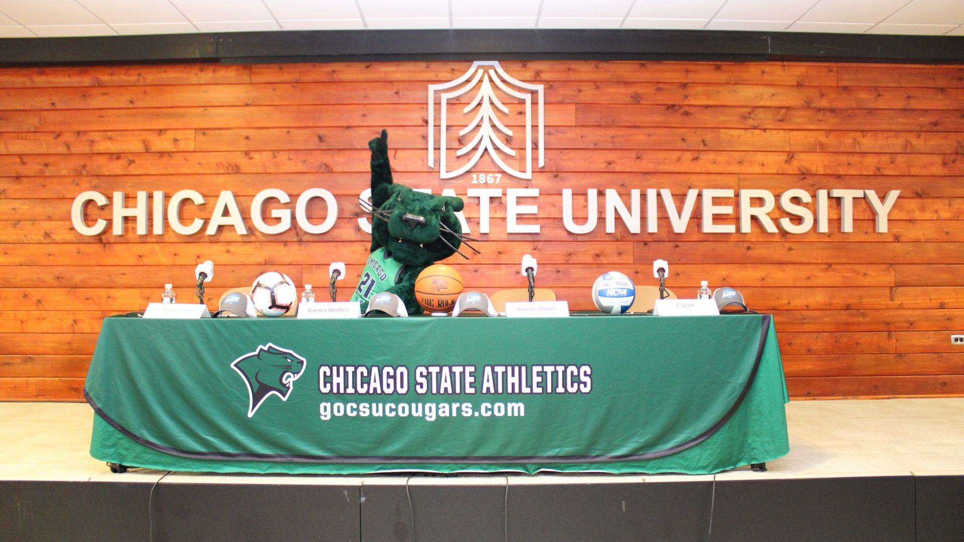 Long table with green tablecloth and cougar mascot behind it in front of Chicago State University sign.