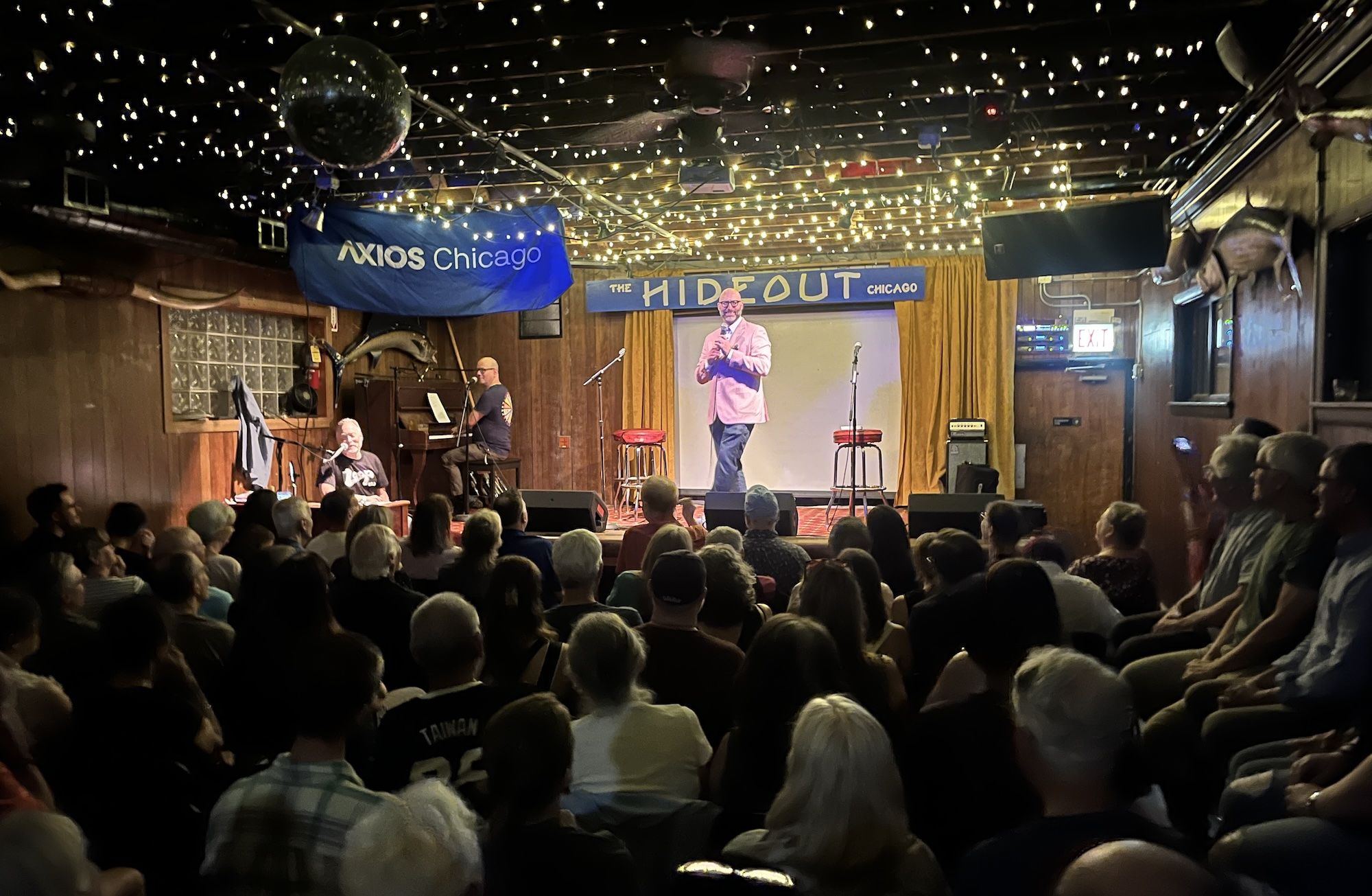 Dimly lit venue with string lights; comedian in a pink blazer on stage at The Hideout Chicago, microphone in hand, audience seated, piano player to the left.