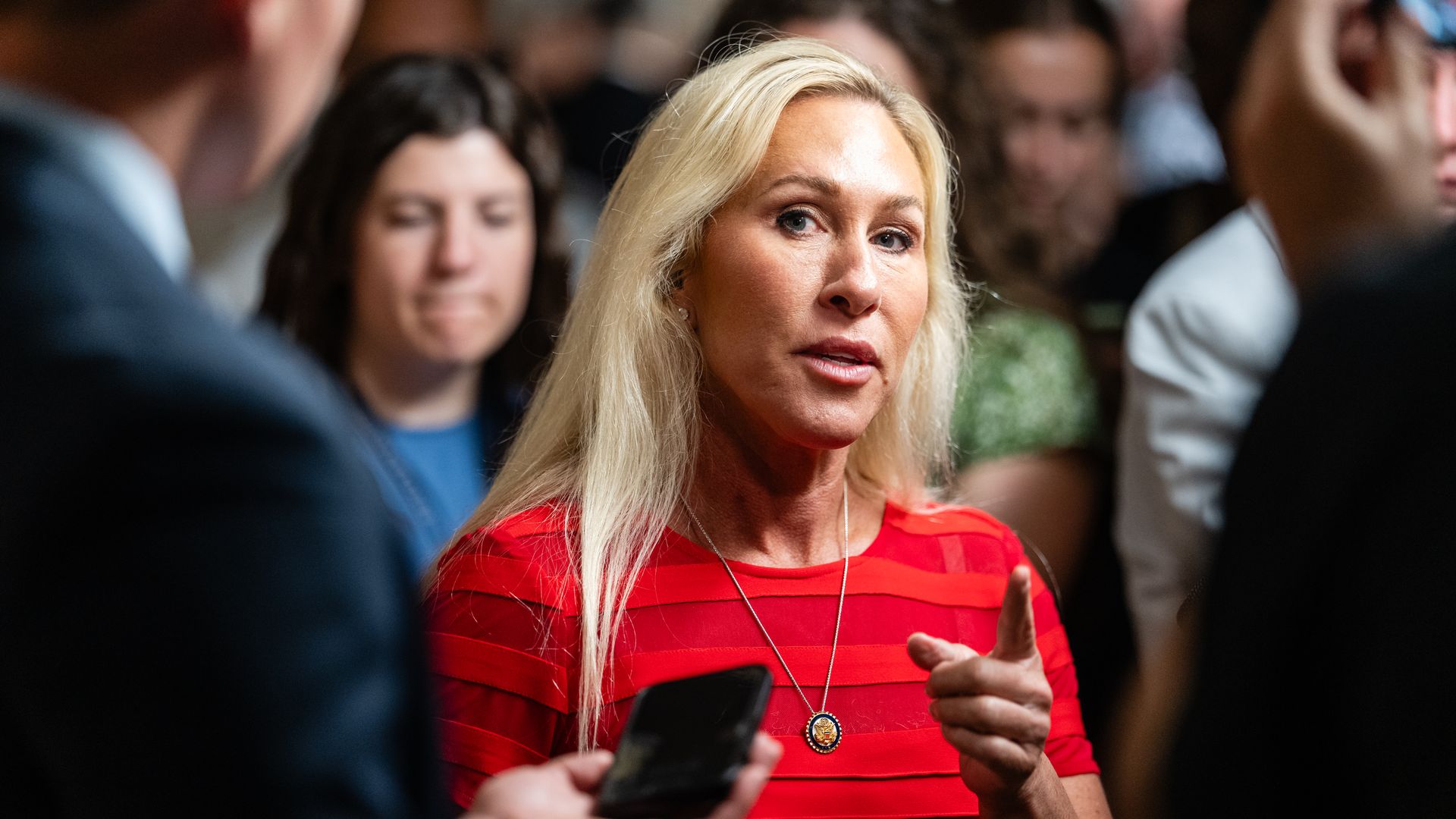 Blonde woman in a red striped top and a pendant necklace speaks emphatically, pointing her finger, surrounded by blurred people in a crowded indoor setting.