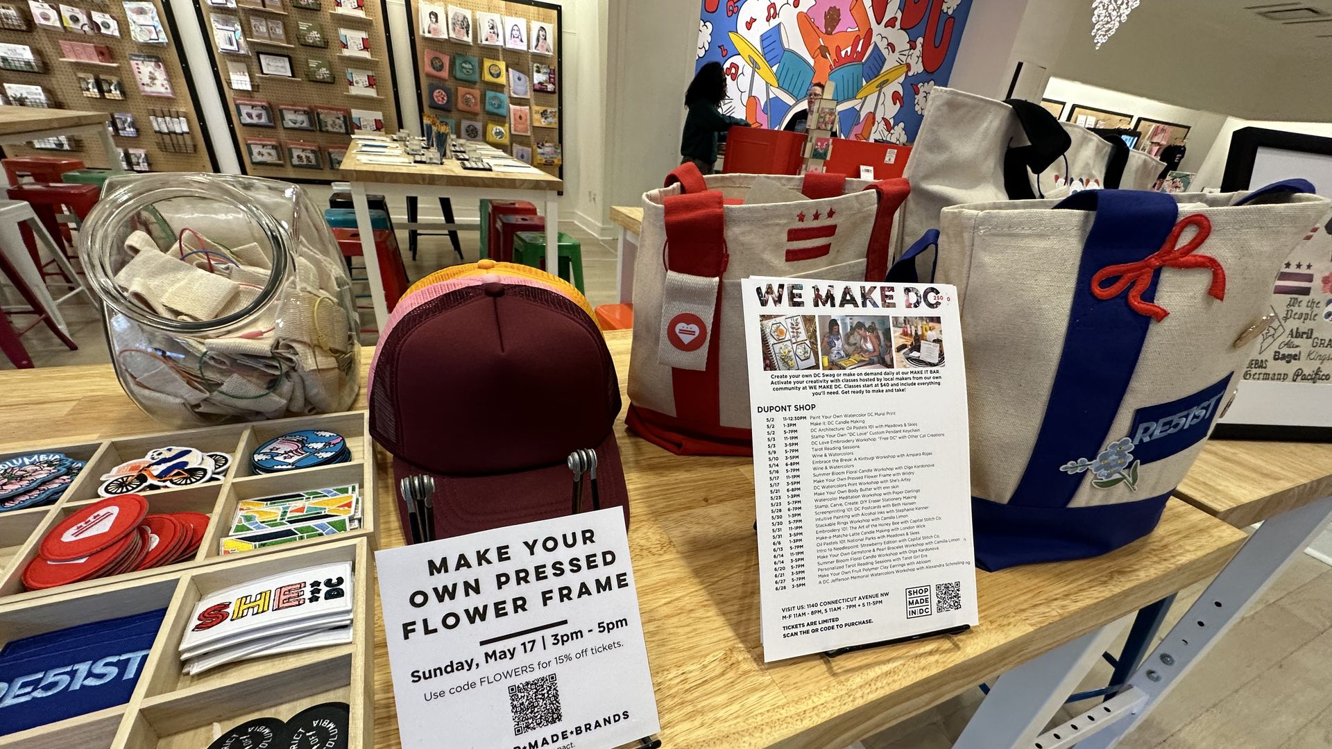 Interior of a colorful shop: foreground shows tote bags with red handles, a glass jar of fabric scraps, and small circular patches; background has display boards, a wooden table, and a vivid mural.