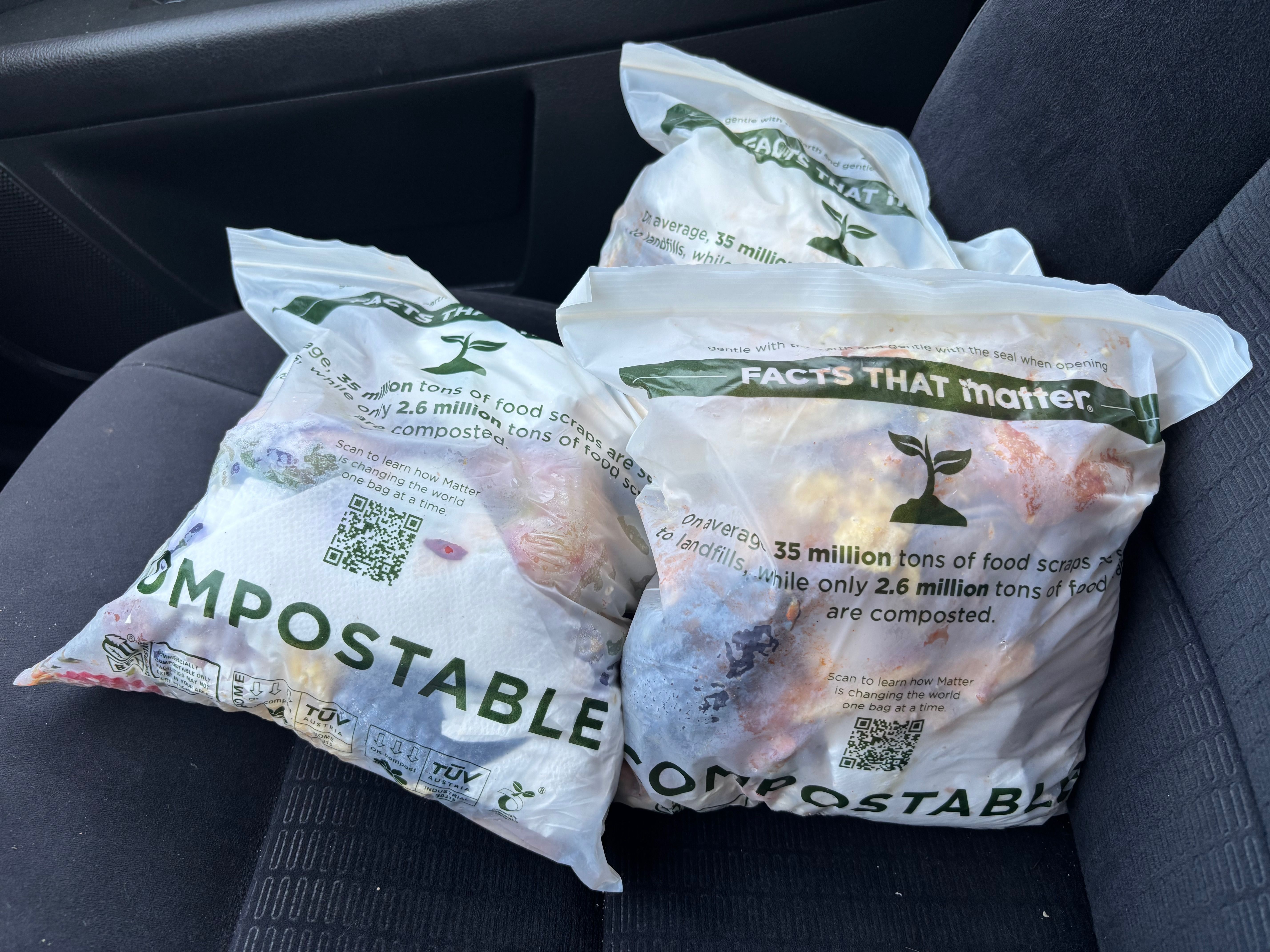 Inside a car, several crumpled white compostable bags with green text and a plant logo lie on the dark seat; labels read "FACTS THAT MATTER" and they contain food scraps.