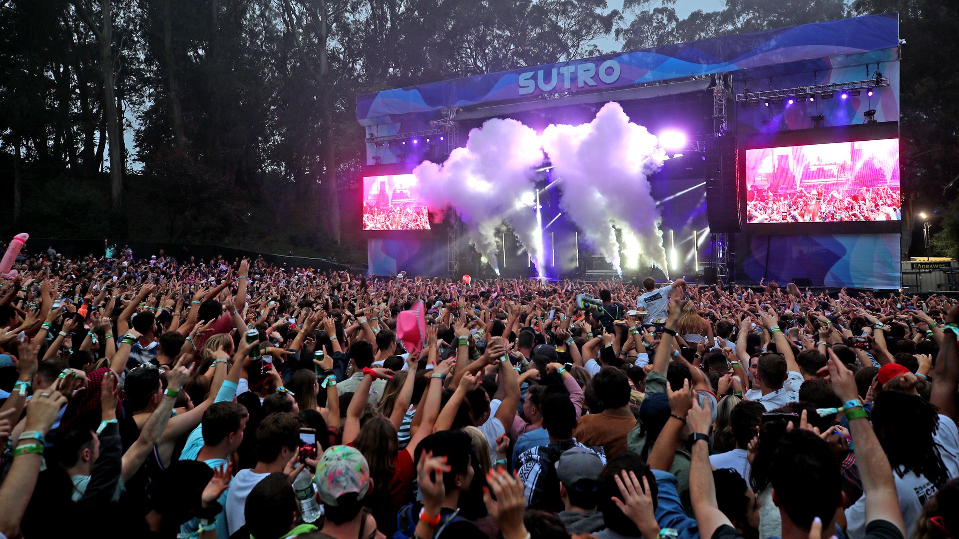 Large crowd at an outdoor concert with hands raised, colorful wristbands, and smoke effects on stage labeled "SUTRO" under evening sky surrounded by tall trees.