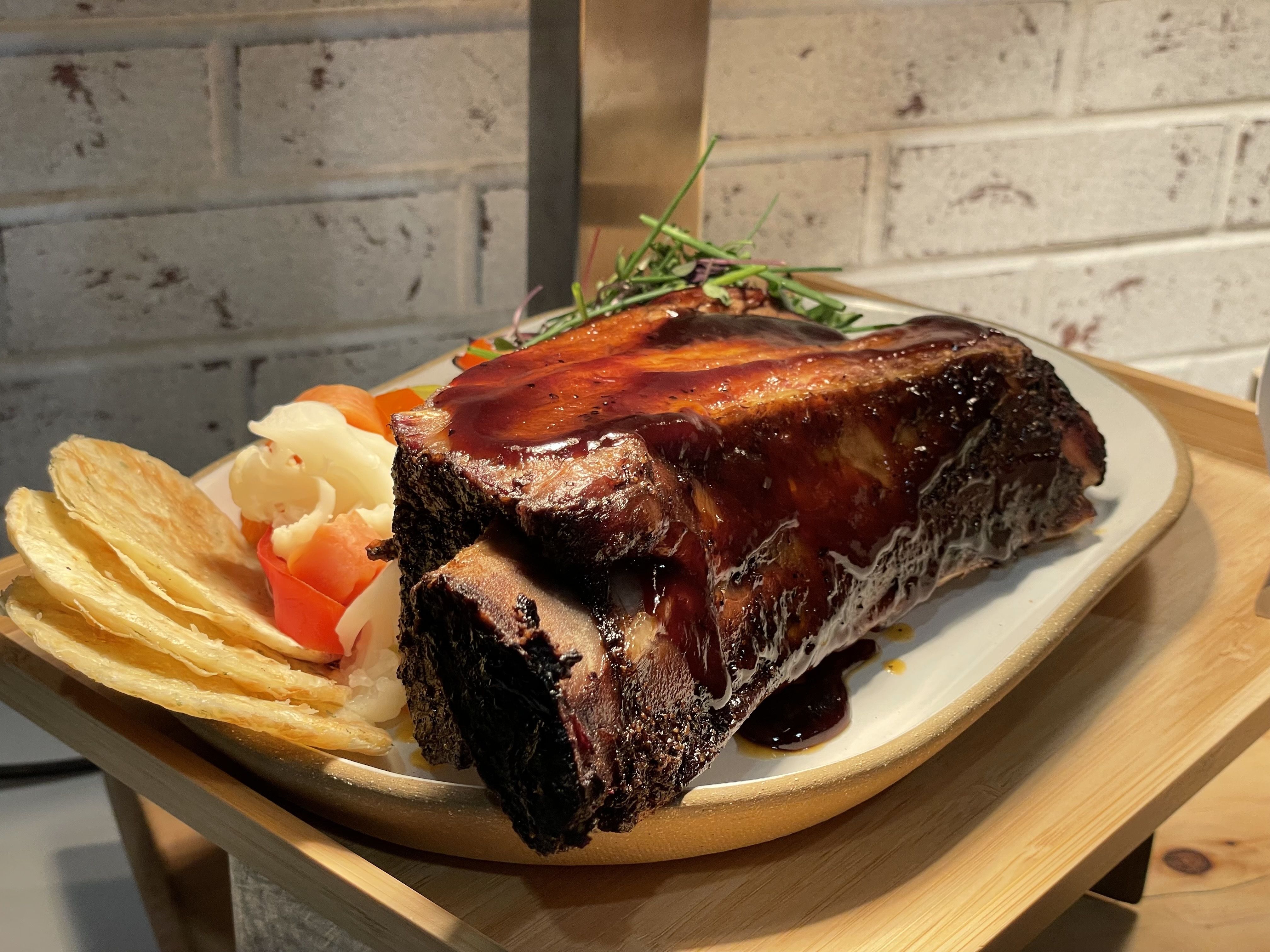 A slab of meat still on the bone, lathered with sauce, on a plate next to potato chops and some veggies. The entire plate is under a bright light. 