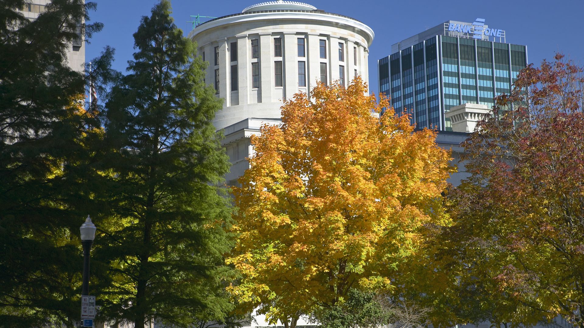 The Ohio Statehouse and a bank tower seen behind autumn trees.