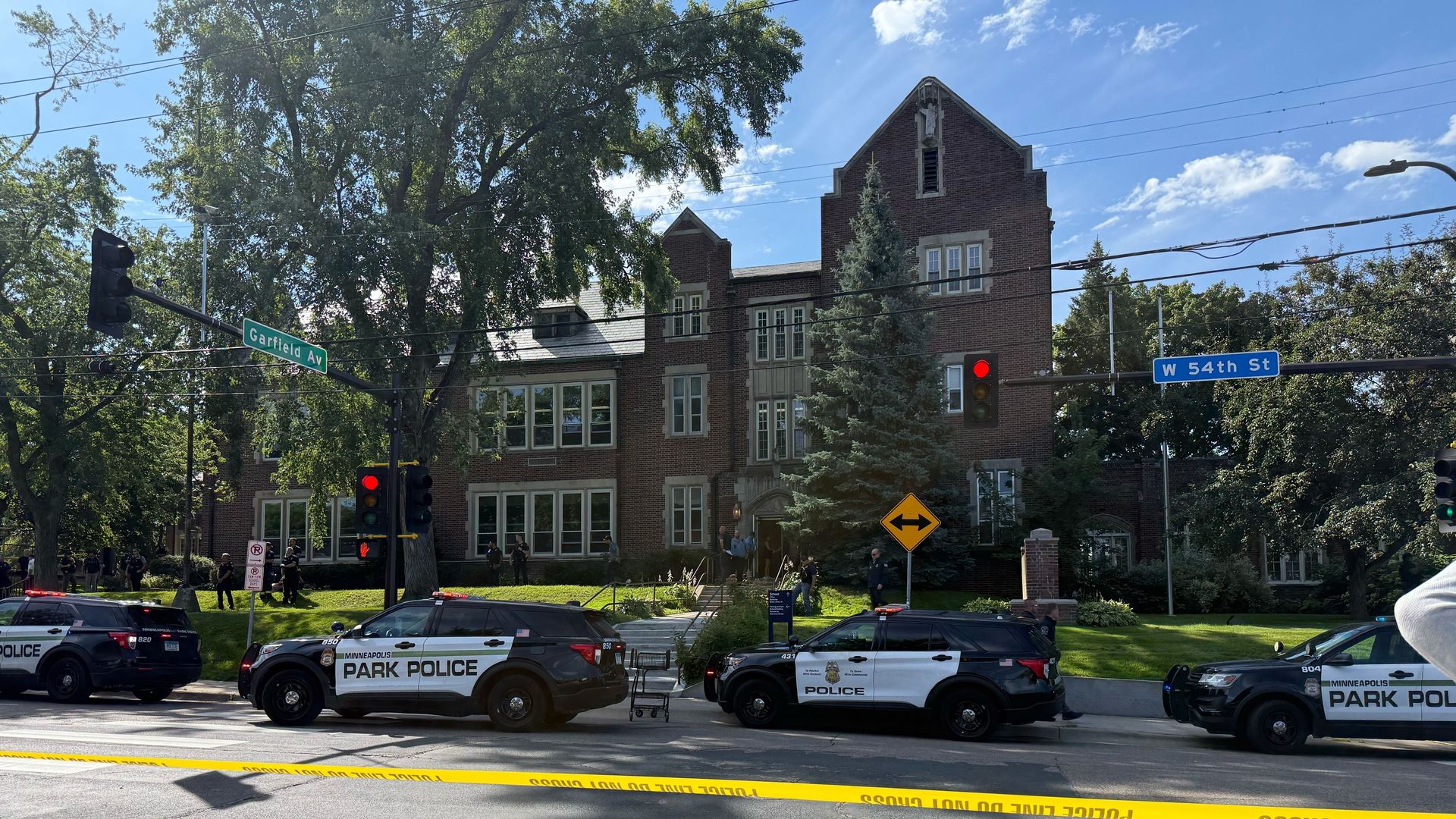 Minneapolis Park Police vehicles block street intersection near a brick building with large trees, yellow police tape across road, and street signs for Garfield Ave and W 54th St on a sunny day.