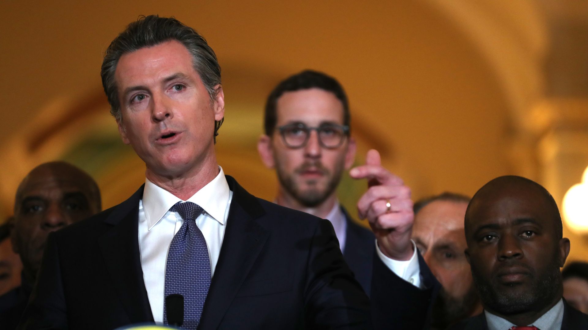 In this image, Gavin Newsom talks and points in a suit with a crowd behind him.
