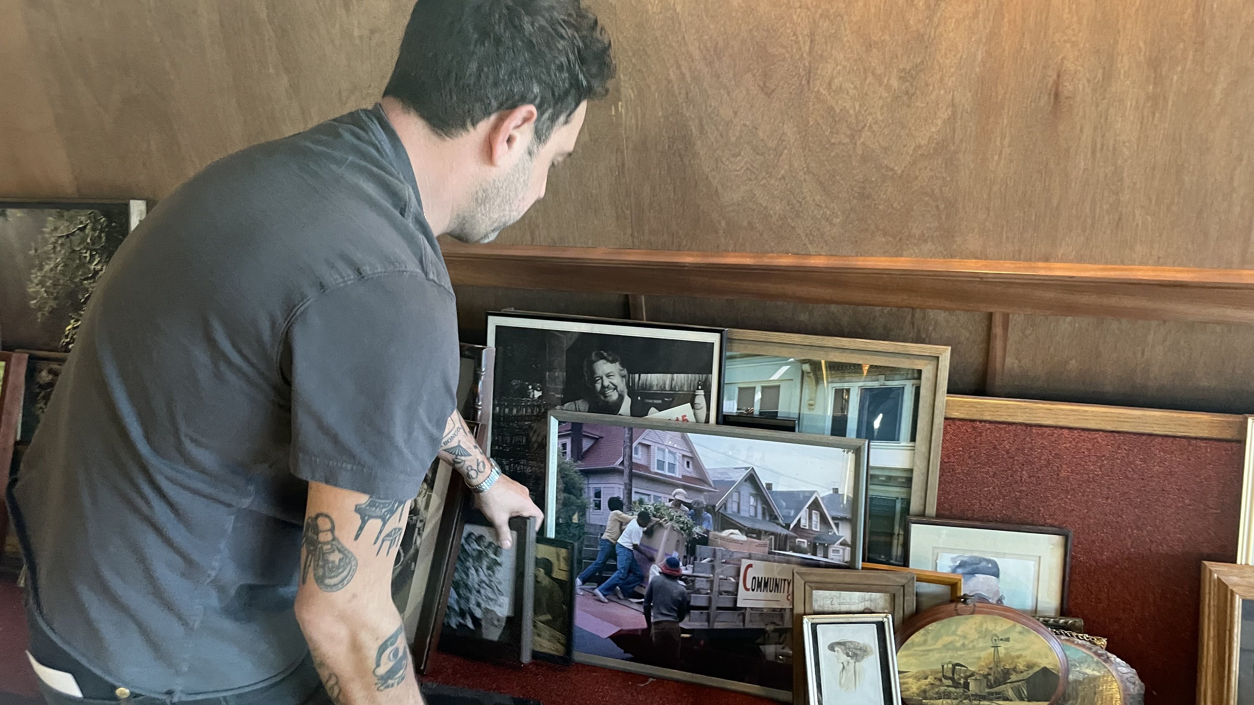A man with tattoos on his arms arranges several framed pictures and paintings on a red carpeted floor against a wooden wall in a cozy room.