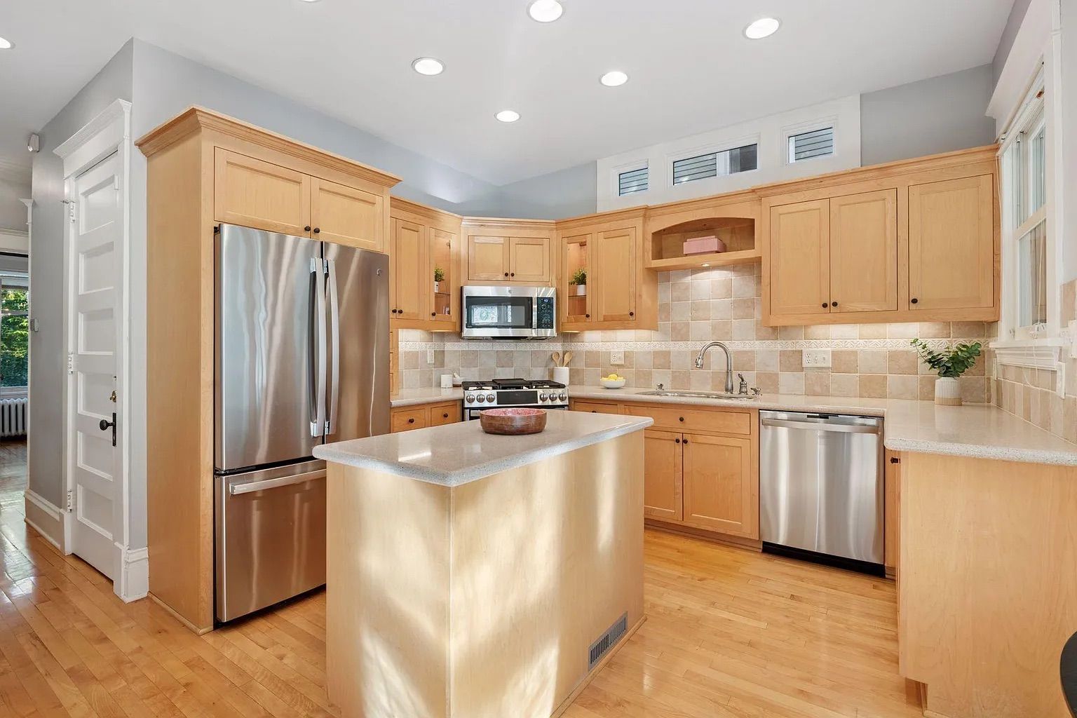Bright kitchen with light wood cabinets, stainless steel refrigerator, oven, microwave, dishwasher, center island, tiled backsplash, hardwood floors, and recessed ceiling lights.