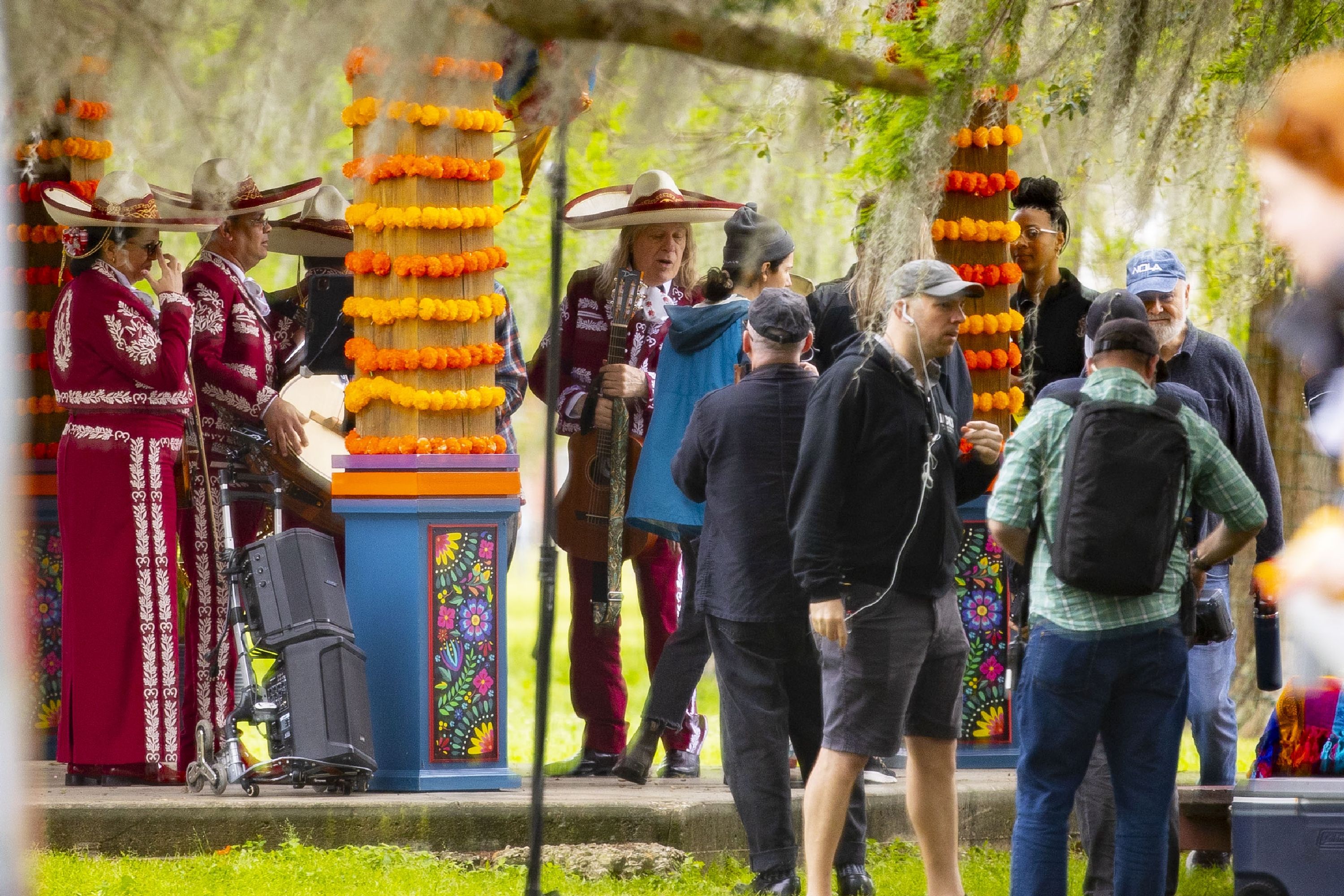 A vibrant outdoor scene with a group of people gathered, some dressed in ornate maroon and white costumes with wide-brimmed hats, resembling traditional mariachi outfits. They stand near colorful pillars decorated with orange marigold garlands. 