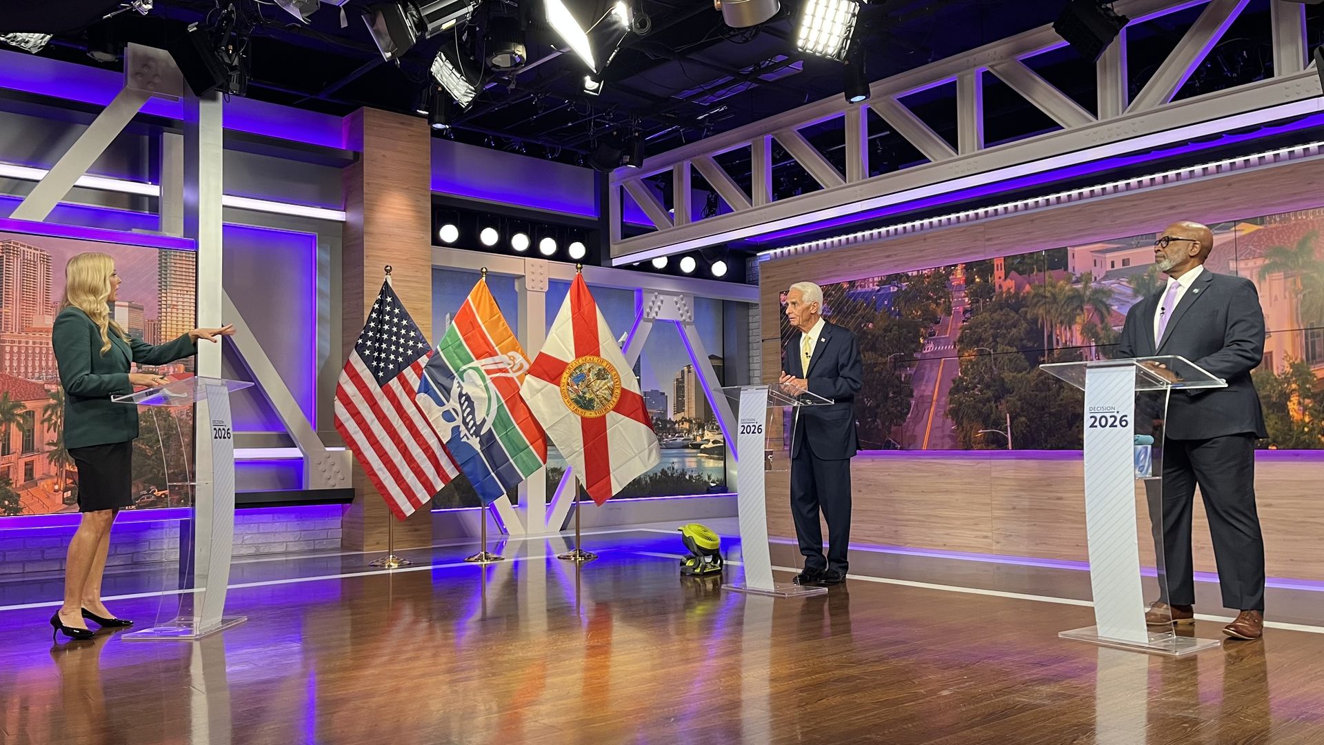 Three-people debate on a TV set: a blonde woman at left podium speaks while two suited men stand at right. U.S. and other flags are between them; purple lighting and a cityscape backdrop.
