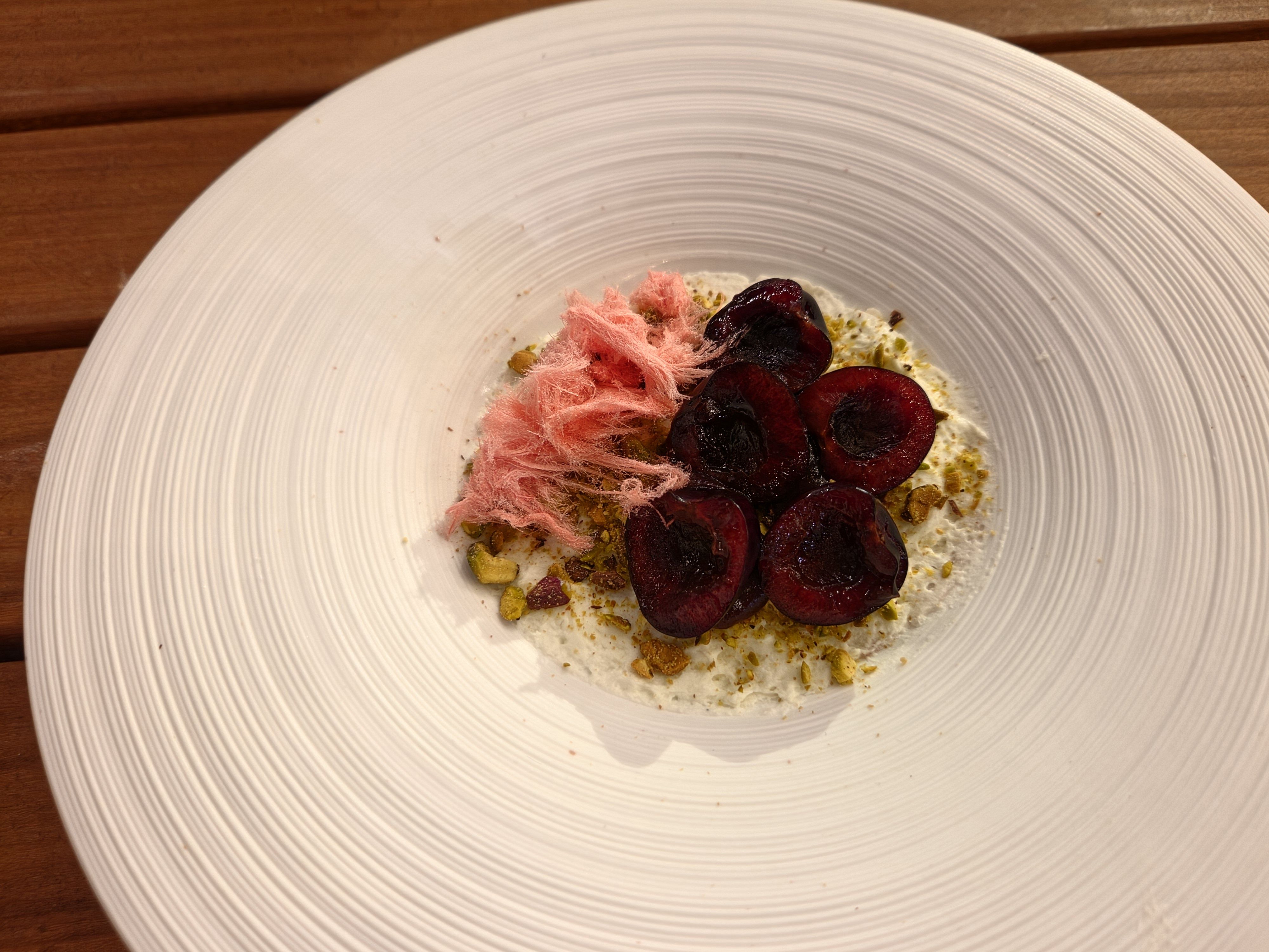 White, ribbed plate on a wooden table, featuring a creamy base topped with dark red beet slices, crushed pistachios, and pink shredded garnish for a plated dessert.