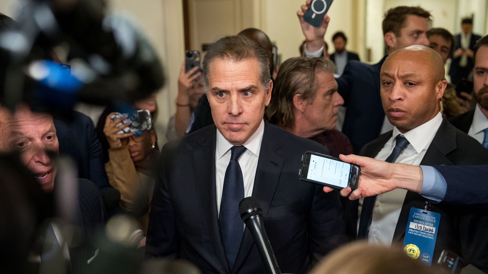 Hunter Biden walks a congressional hall as several people surround him, extending cameras and microphones into his face. 