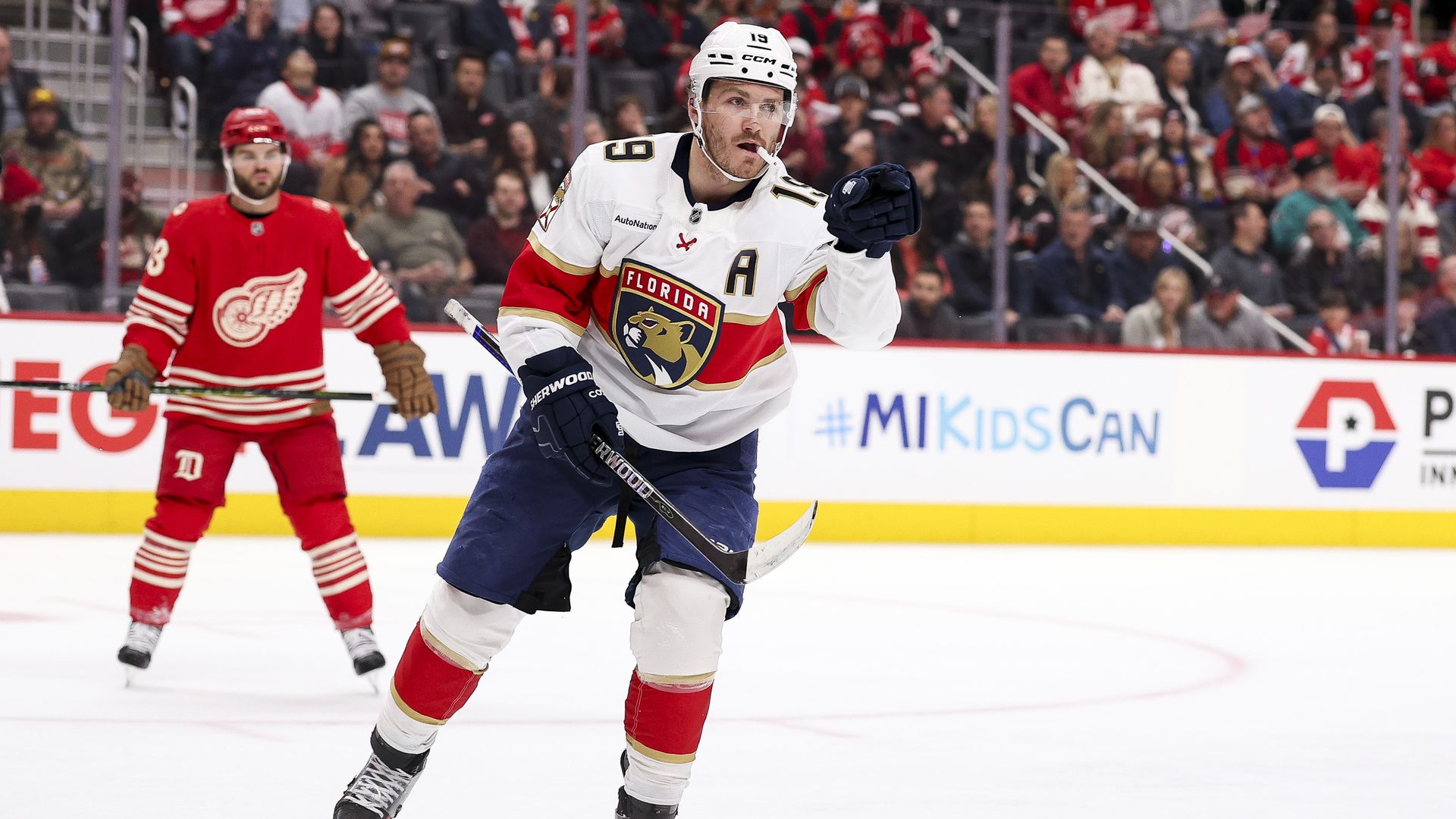 Matthew Tkachuk wearing his white panthers jersey pointing with his mouth guard out of the side of his mouth. A Red Wings player stands in behind on his left in the distance.
