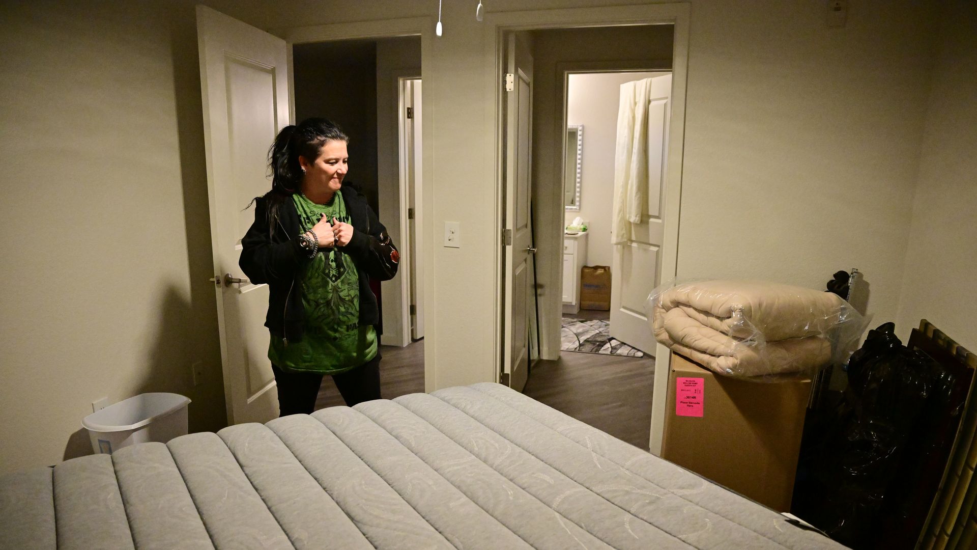 A woman in a green shirt and black jacket stands in a modest bedroom with a gray mattress, beige folded bedding on a box, and open doors leading to other rooms.