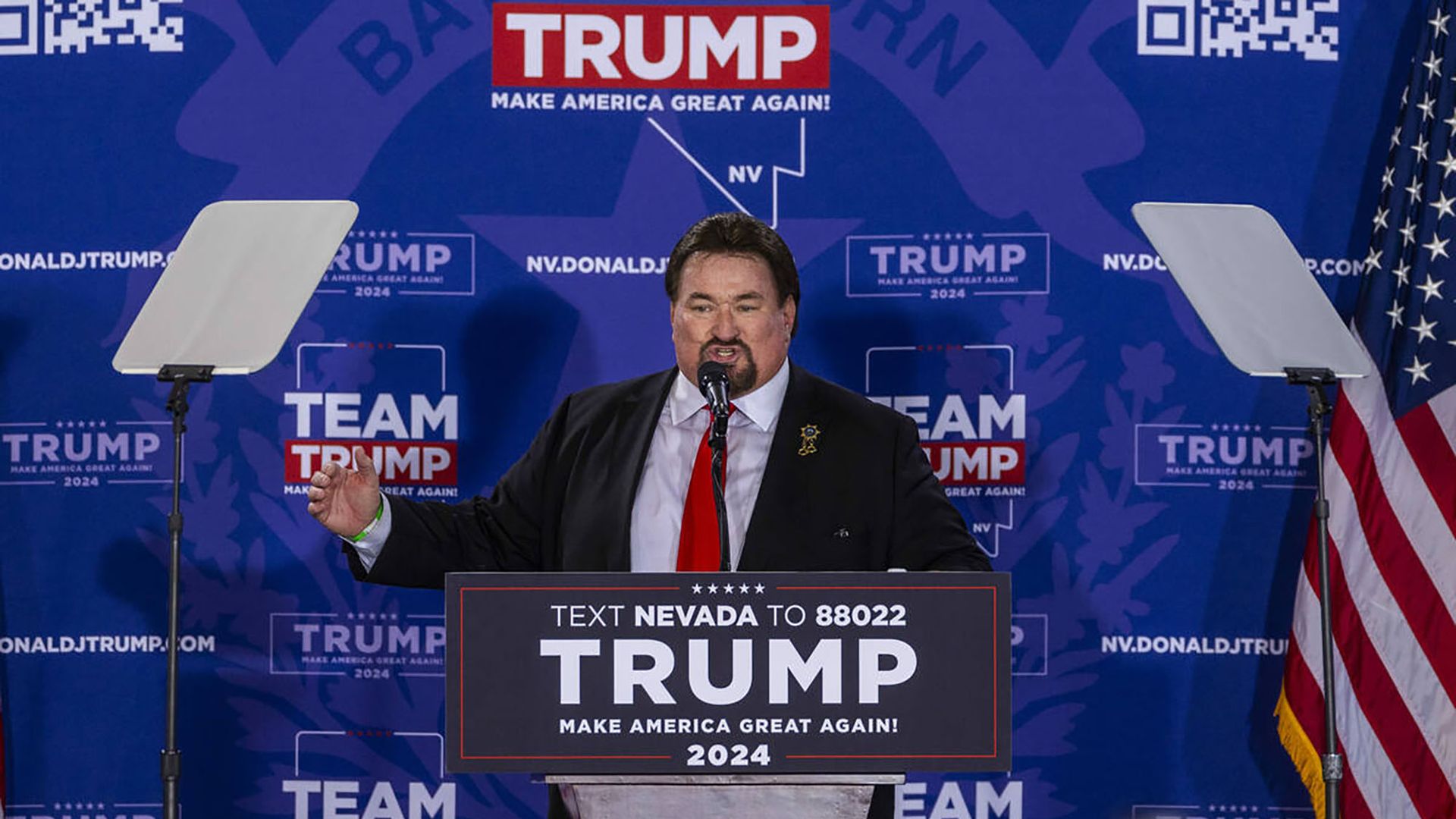 Michael McDonald speaks at a lectern that promotes Donald Trump. The backdrop is also Trump branded. 