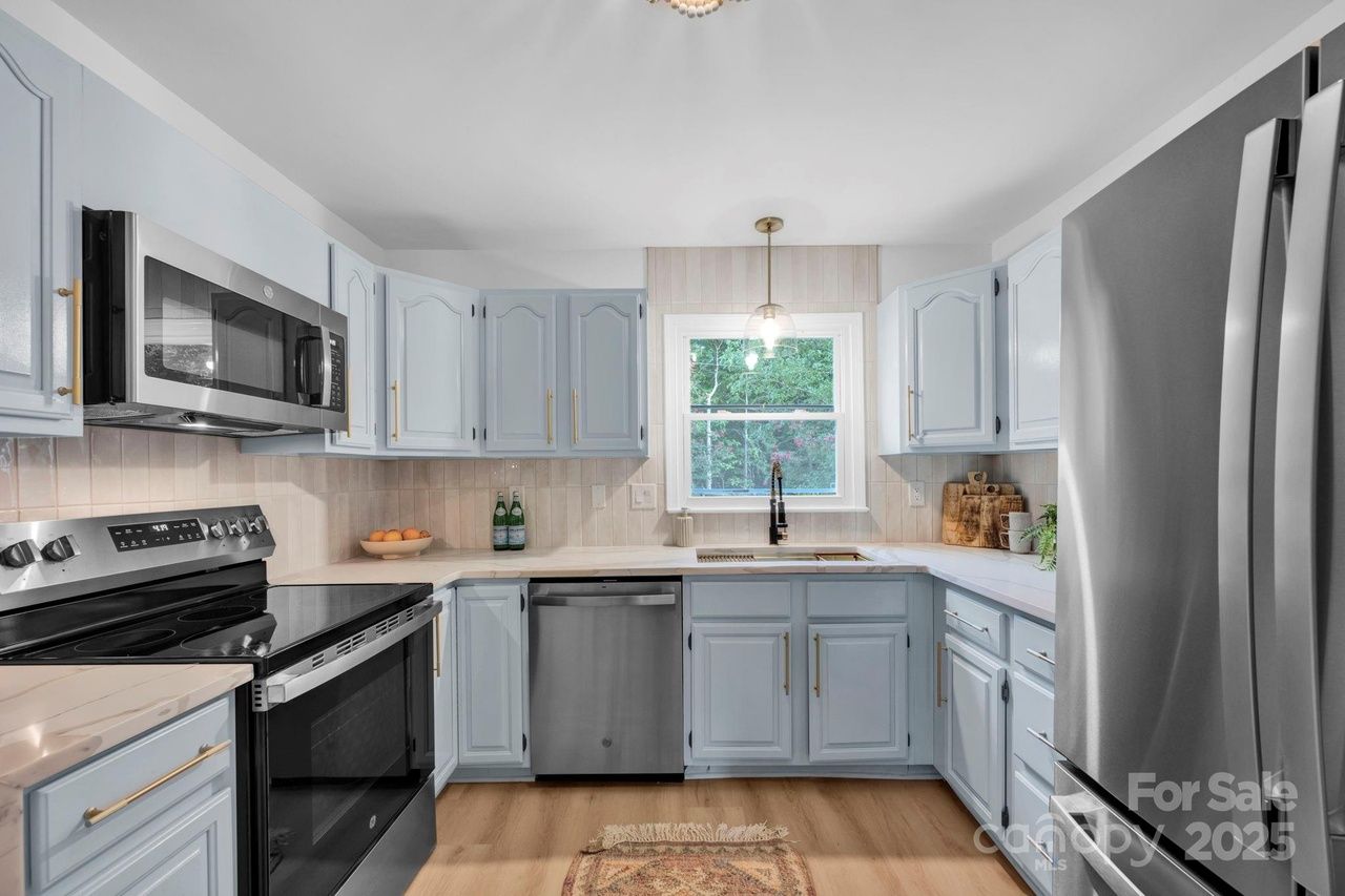 Modern kitchen with light blue cabinets, stainless steel appliances, white countertops, wood flooring, a window above the sink, and a small rug on the floor.