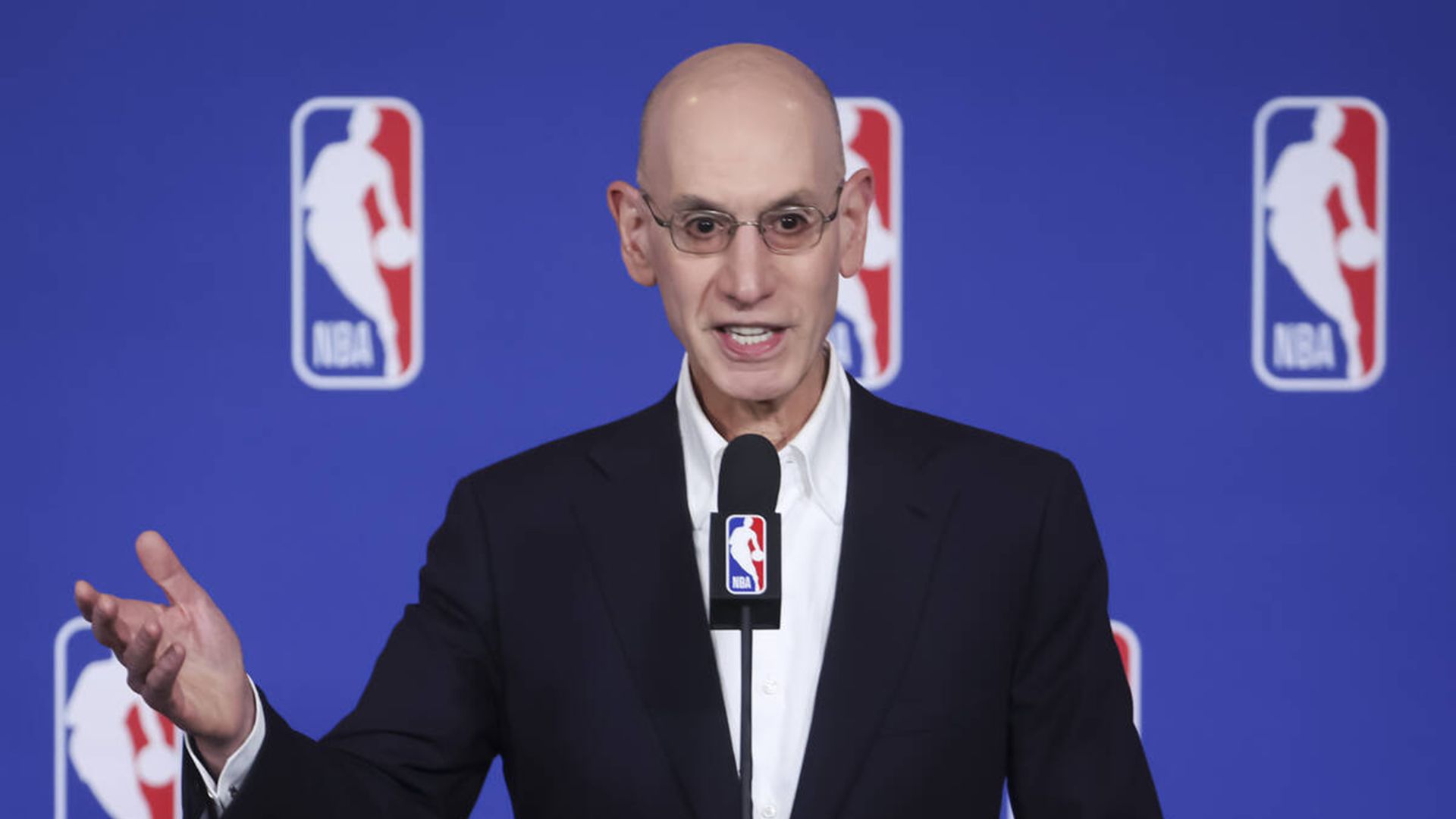 A man wearing glasses in a dark suit speaks at a podium with a microphone, gesturing with his hands, against a blue backdrop with NBA logos.