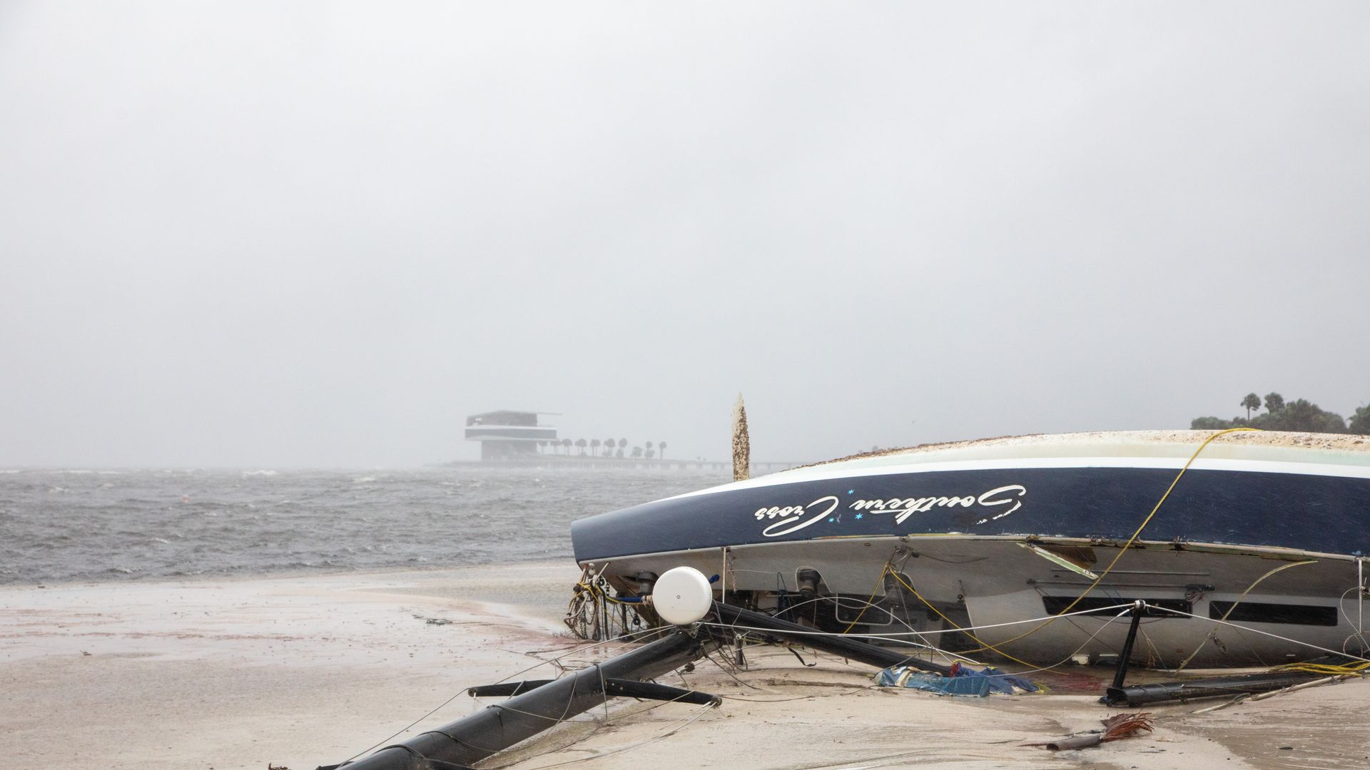A sailboat grounded on a sandy shore and turned on its side with gray water, a gray sky and a pier in the background.