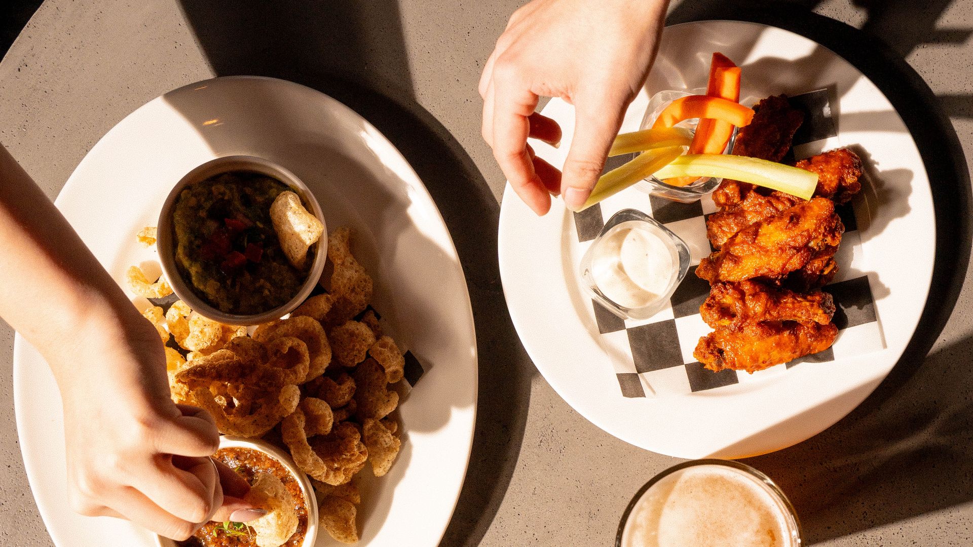 An overhead view of three plates. Two hold chicken wings and dipping sauces with vegetables. A third has fried shrimp. Three hands are reaching for food, one holding a glass of orange juice, at Anaqua Garden Bar.