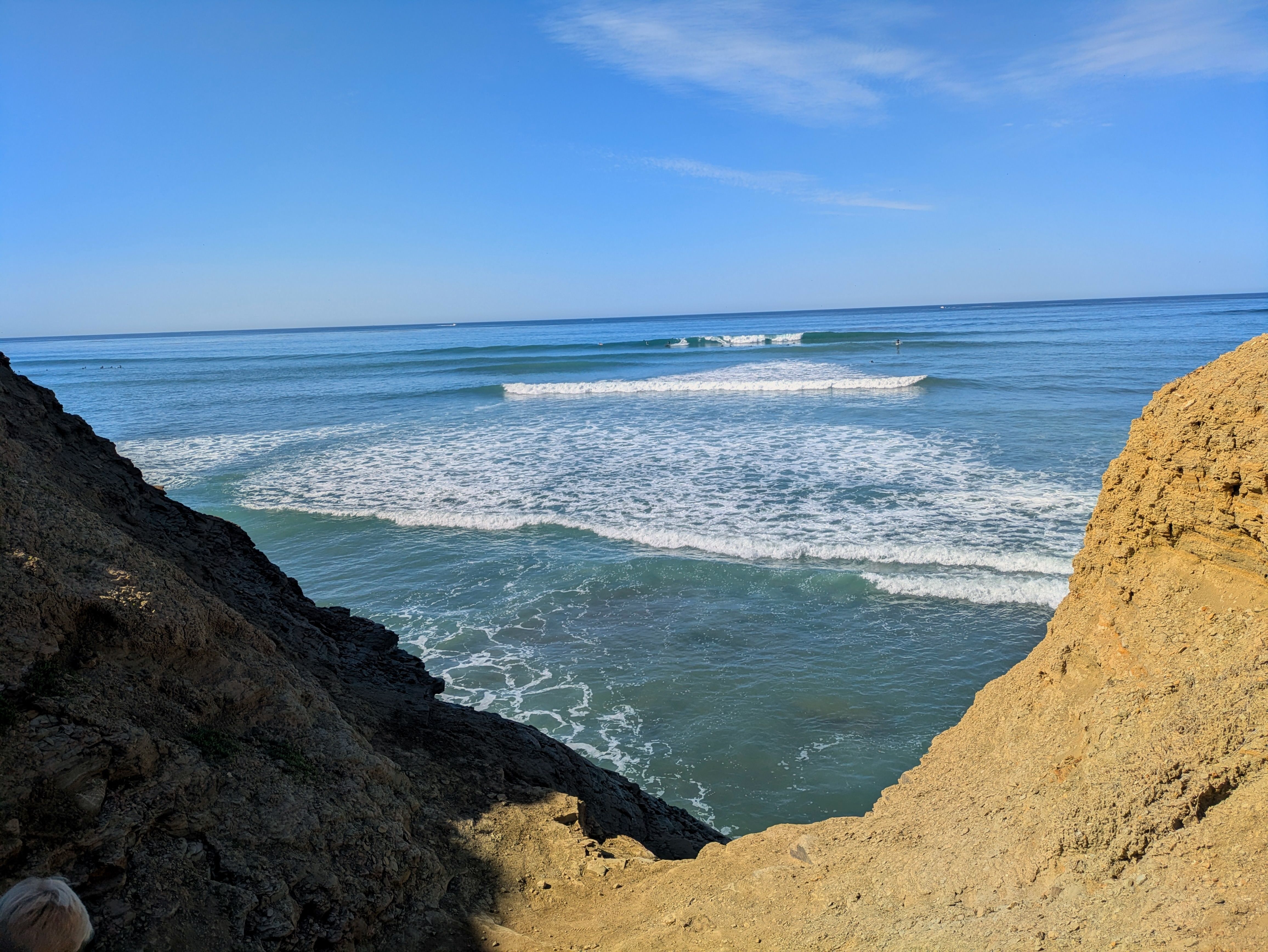 View of ocean waves breaking in lines under a blue sky, framed by rocky brown cliffs on the left and right sides.