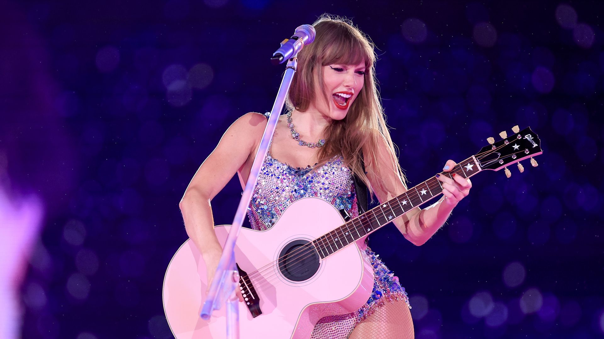 Taylor Swift strums a pink guitar in a sparkly leotard on stage.