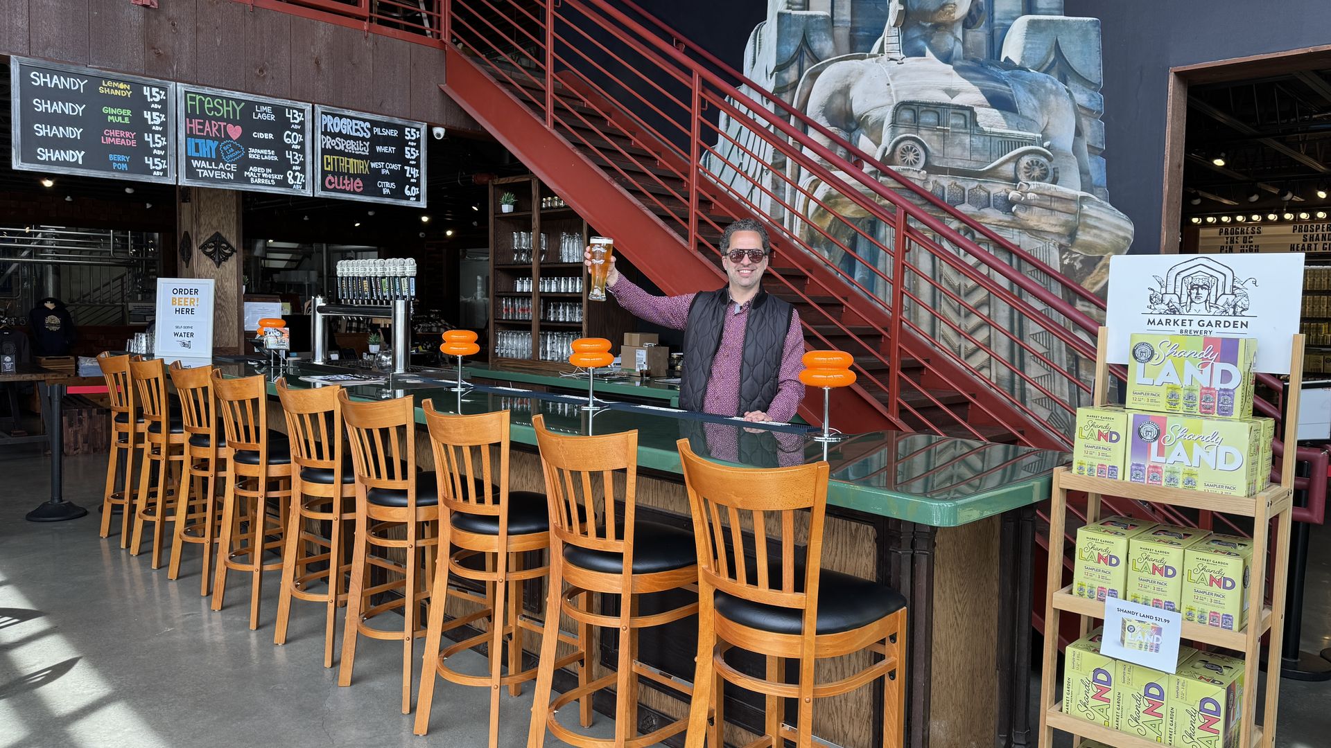 Man in sunglasses raising glass of beer behind green bar in front of red staircase
