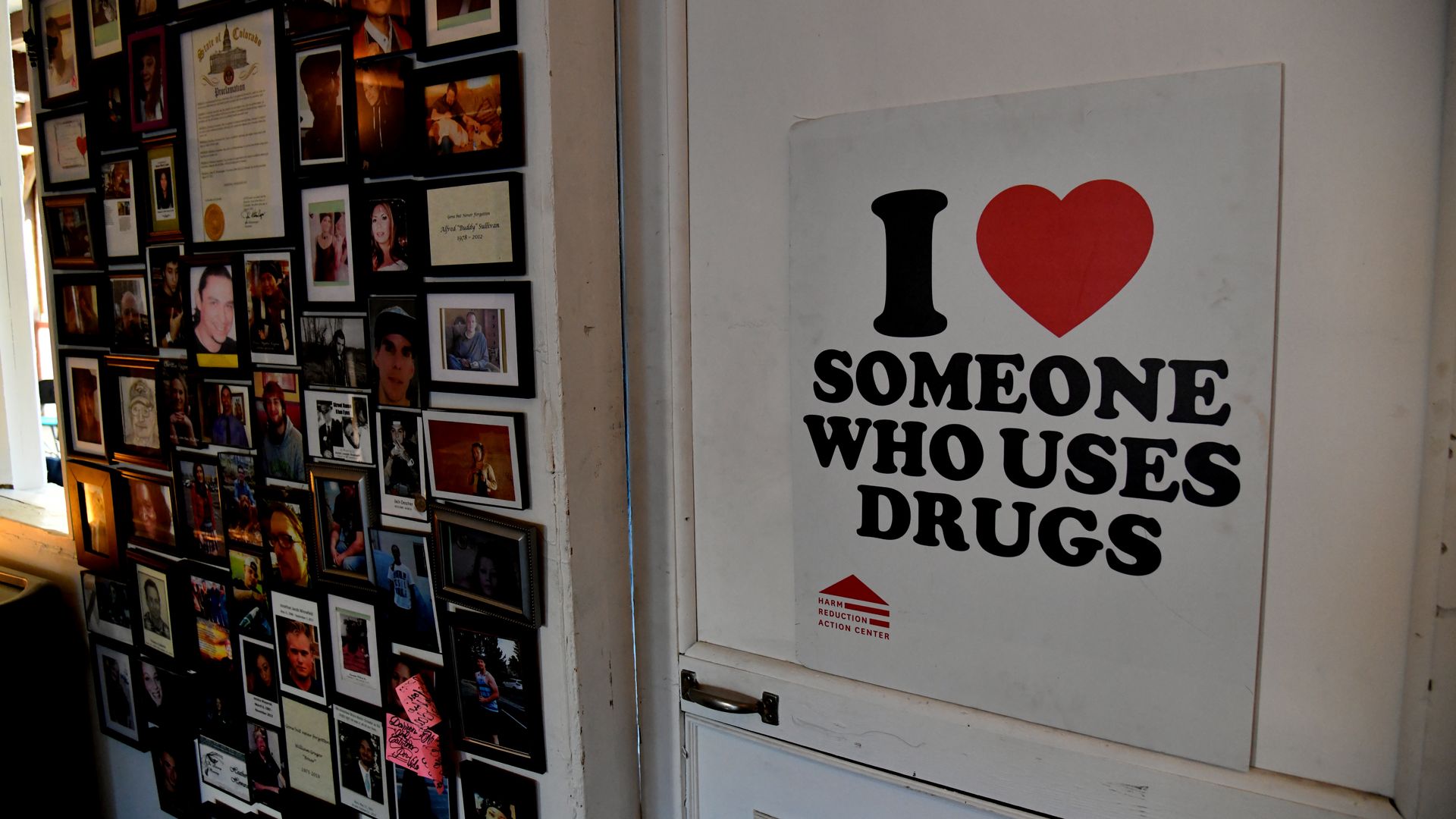 A sign that reads I HEART SOMEONE WHO USES DRUGS next to several framed portraits. 