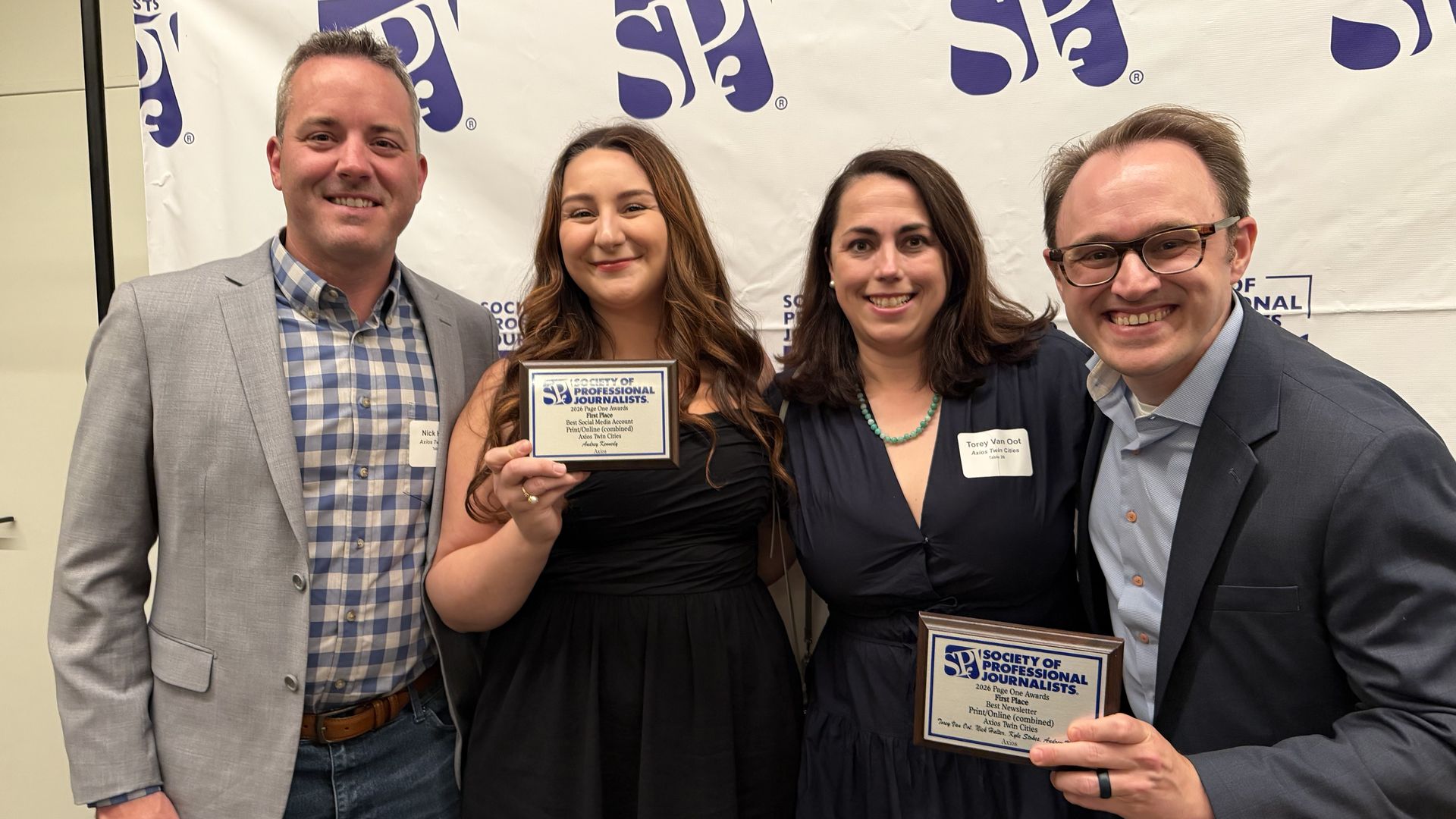 Four people pose at a Society of Professional Journalists event. A woman in a black dress holds a plaque; two men in suits and another man in a checkered shirt smile in front of a SPJ banner.