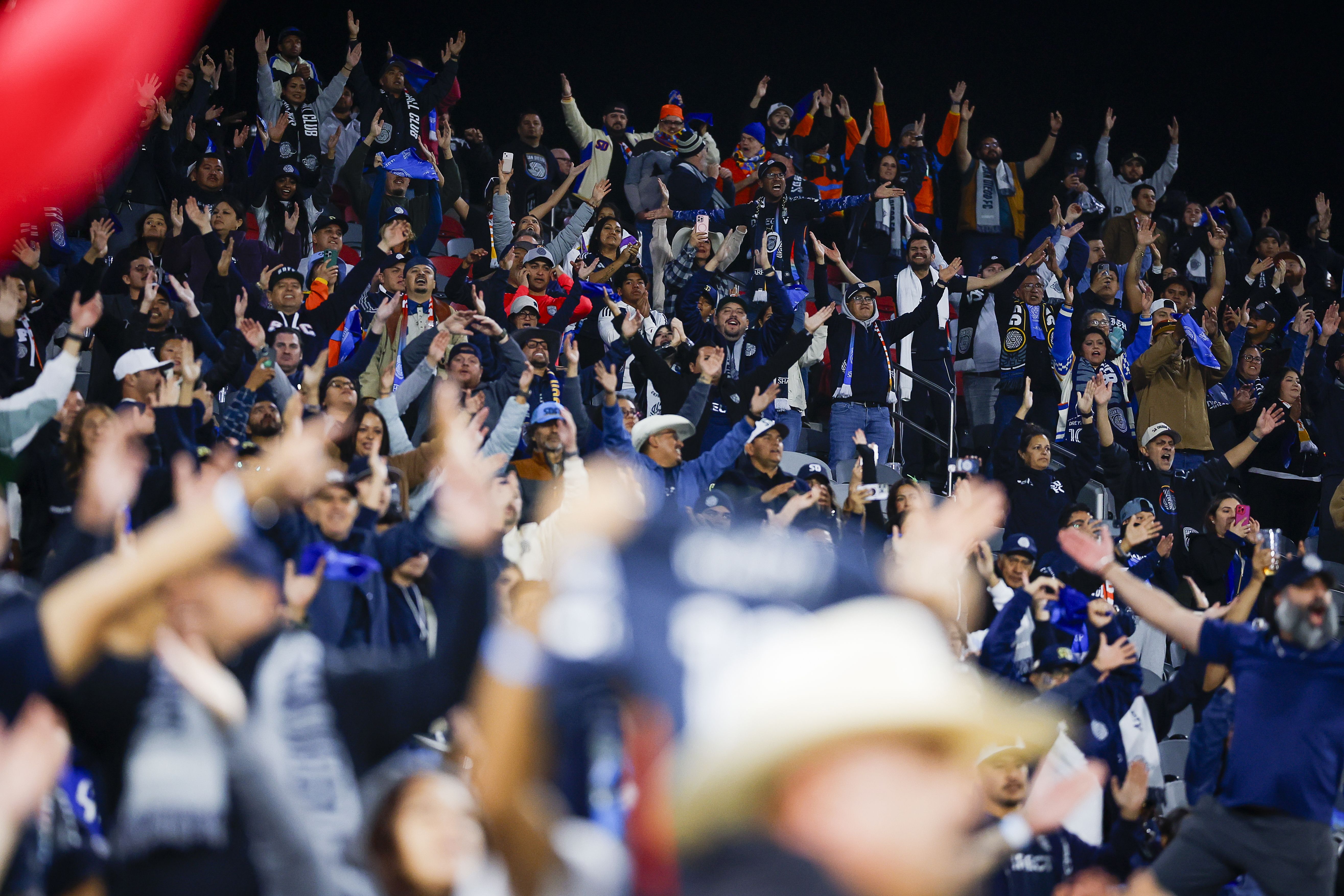 A crowd of San Diego FC fans chant with arms raised in a V to clap in the stands.