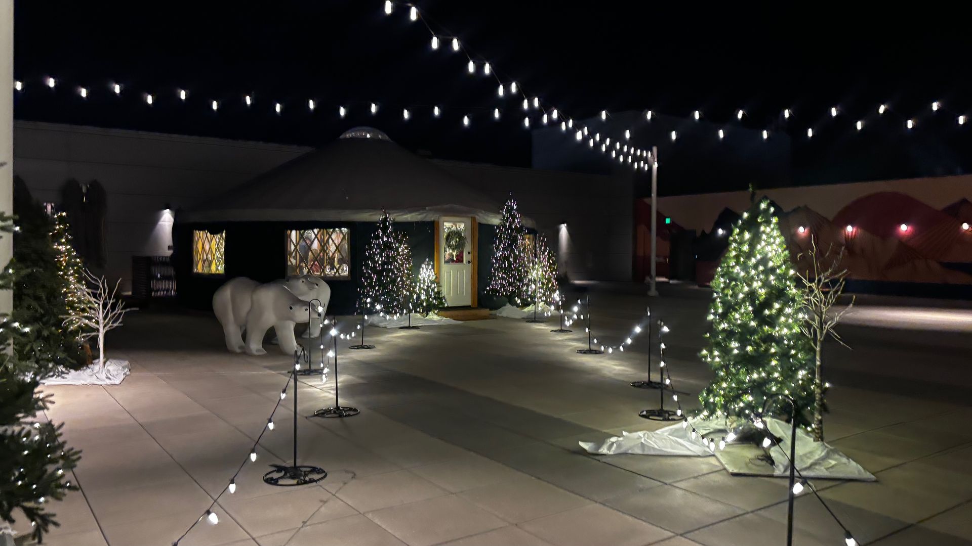 Night scene with Christmas trees lit by white lights, a small yurt decorated with wreath on the door, two polar bear statues, and string lights overhead creating a festive atmosphere.