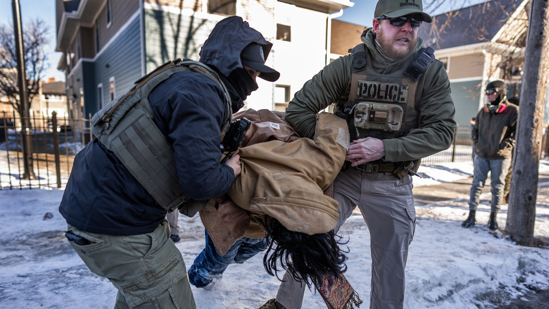 Two officers in tactical gear lift a person on a snow-covered residential street; a helmeted bystander watches in the background.