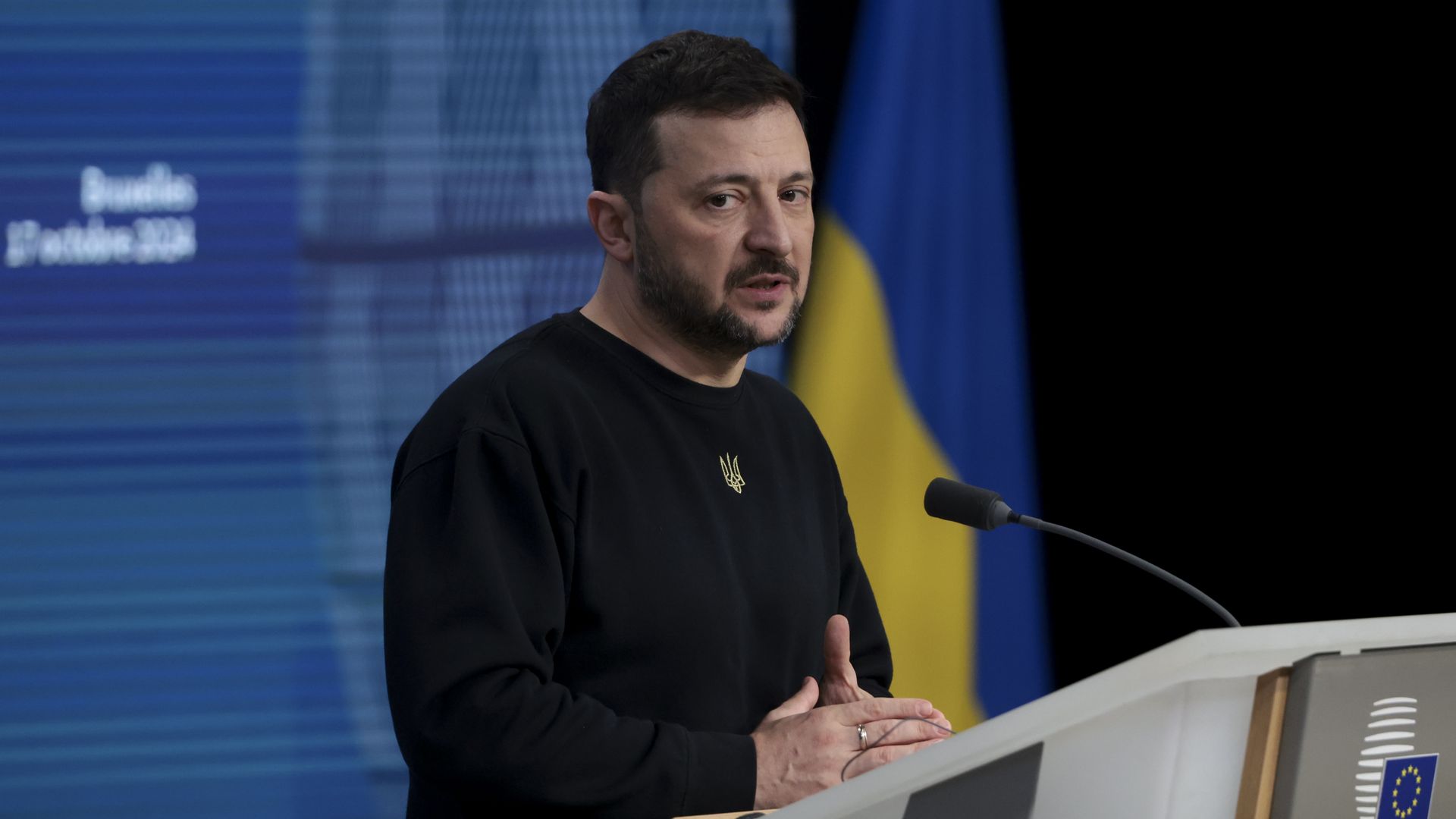 Wearing a black shirt, Zelensky speaks into a microphone at a lectern. 
