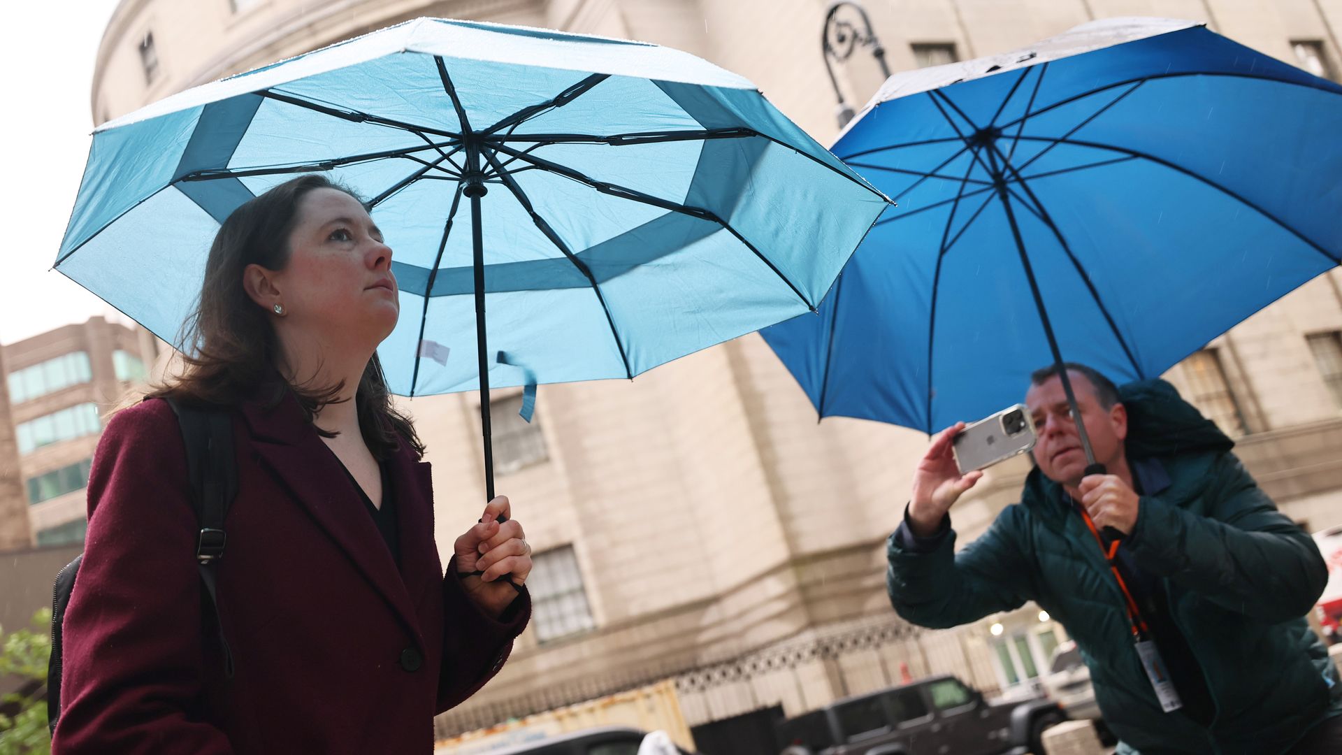 A woman with brown hair wearing a red coat (Maurene Comey) carries a blue umbrella while a man in a green jacket, also carrying a blue umbrella, appears to take a photo of her with his iPhone.