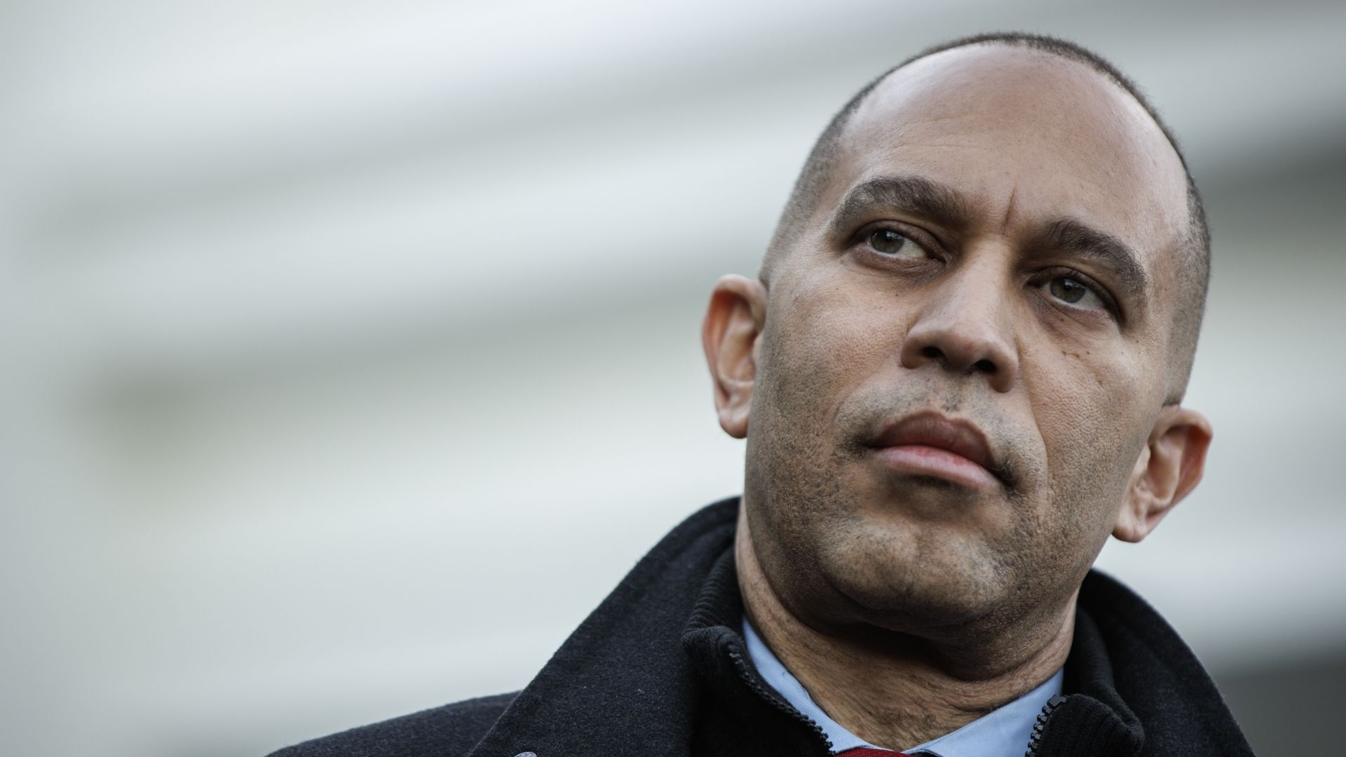 Hakeem Jeffries, wearing a black jacket and blue shirt, standing in front of a white building.