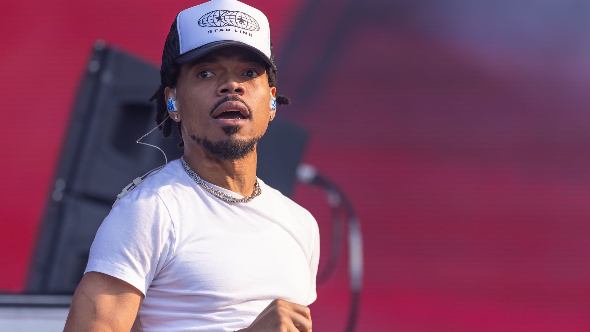 Man with goatee wearing a white T-shirt and a black and white cap with "STAR LINE" logo, holding a microphone on stage with a red background.