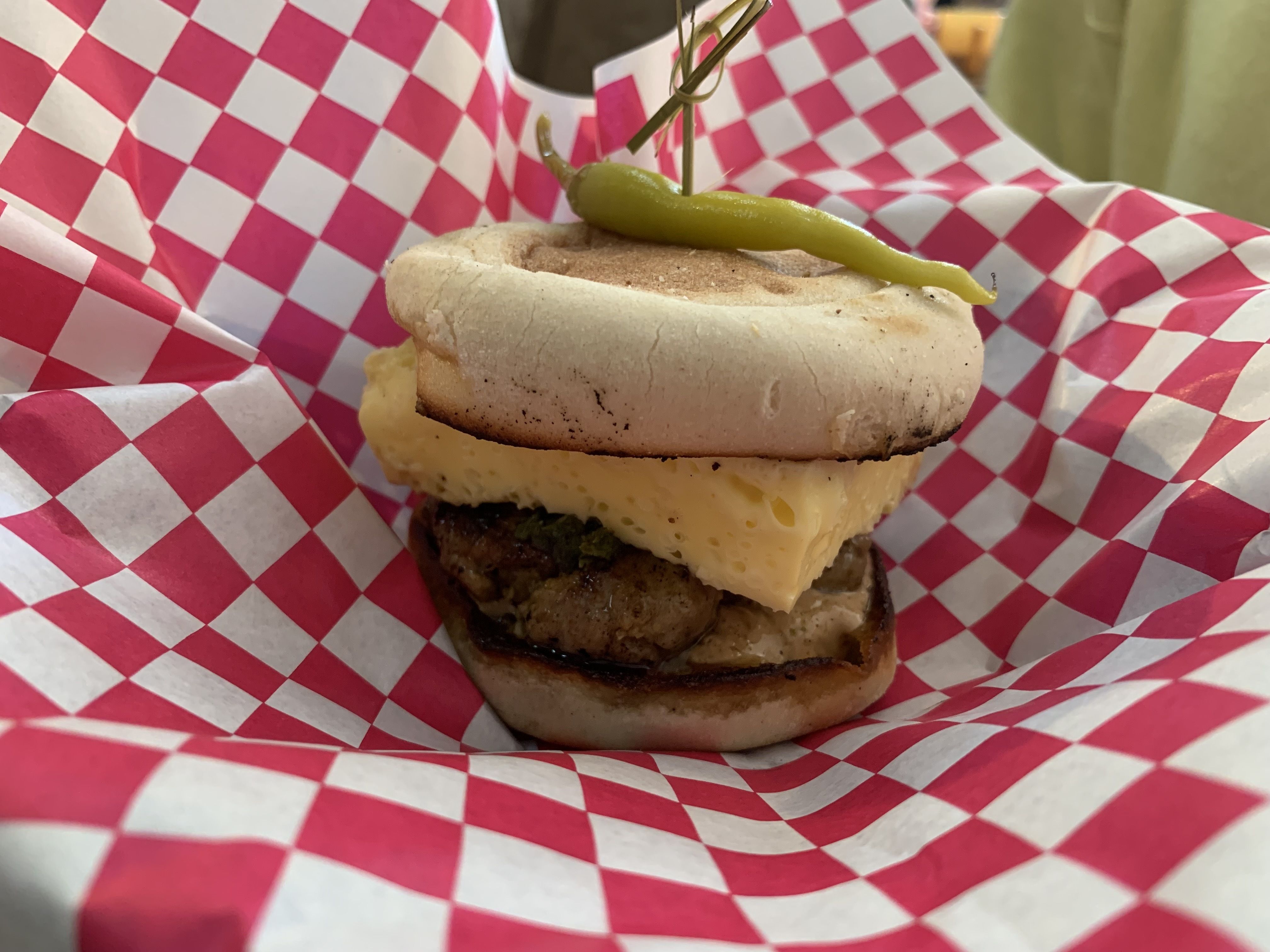 Close-up of a cheeseburger with melted cheese, two beef patties, and a bright green pickle spear skewered on top, all inside a toasted bun, wrapped in pink and white checkered paper.