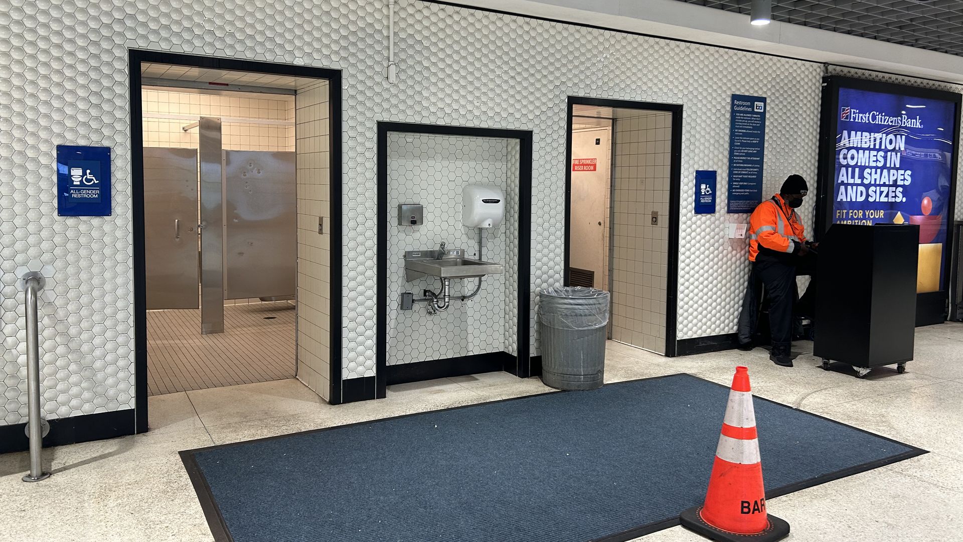 Interior with white hex tile walls and three doorways. Left: open bathroom; middle: sink; right: worker in an orange vest at a kiosk by a bank ad. An orange cone on a blue mat.