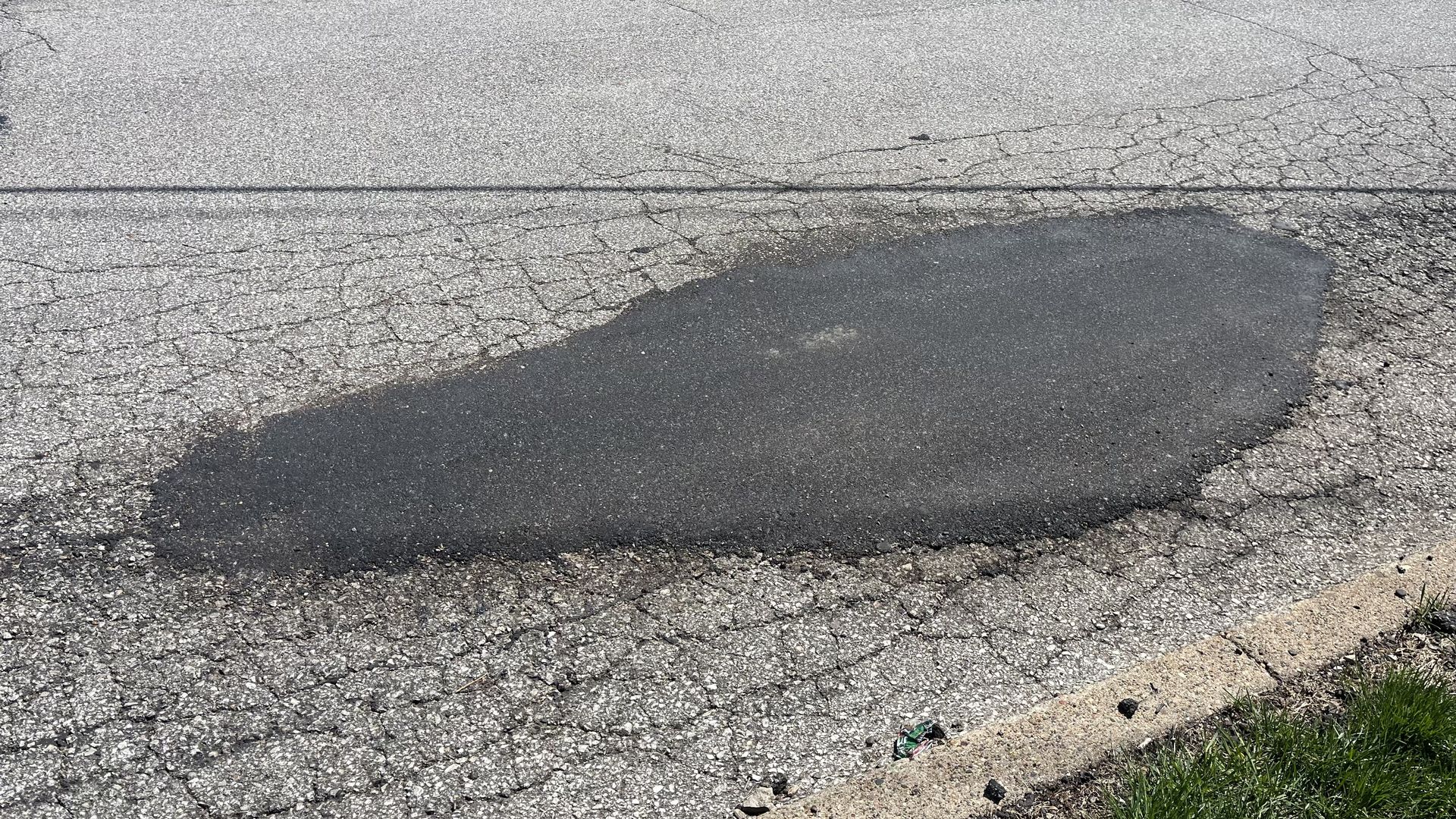 Cracked asphalt road with a large curved patch of fresh black pavement along the lower center, a straight seam across the upper section, and patches of grass at the bottom right.