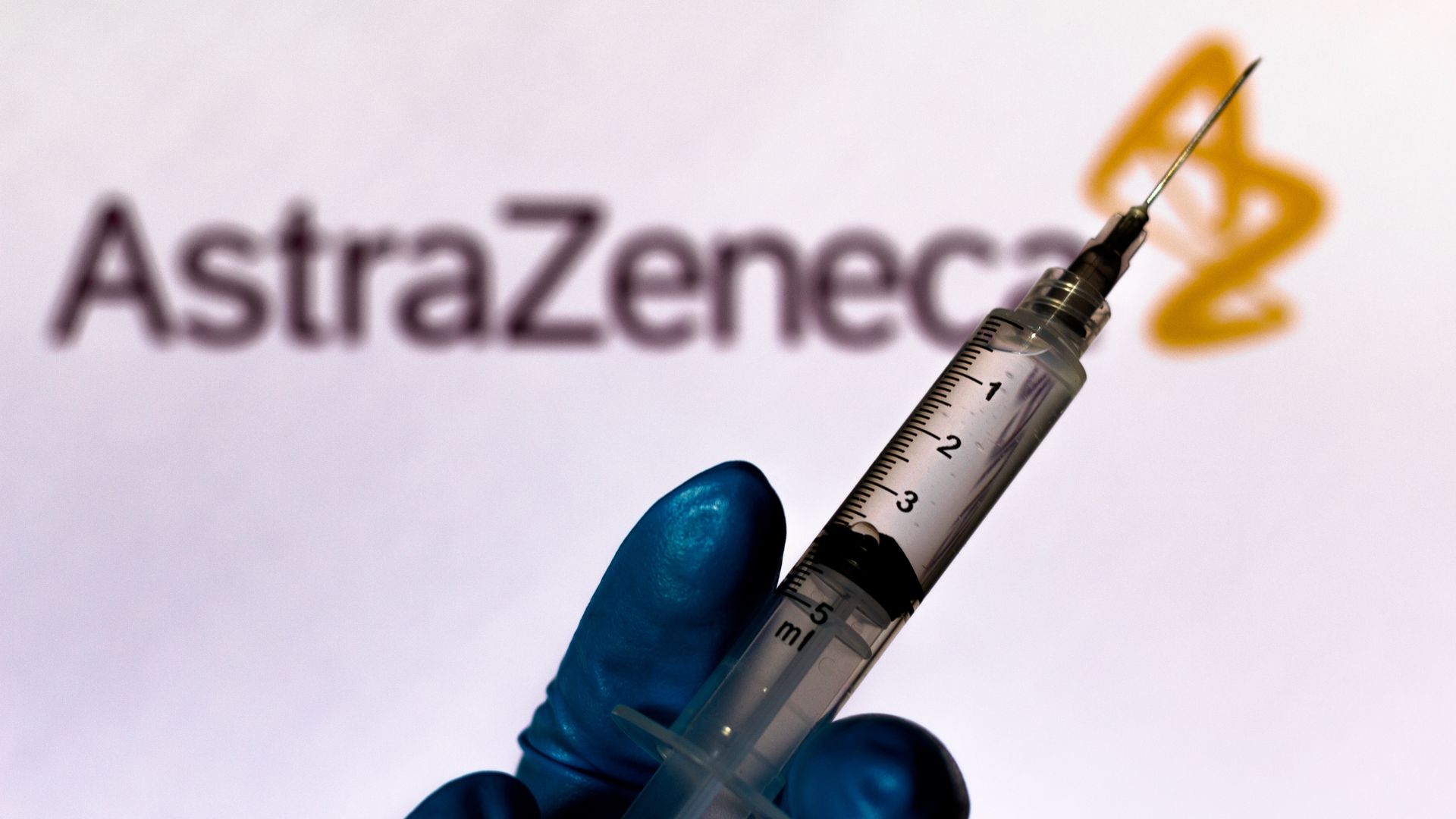 Picture of a gloved hand holding a syringe in front of an AstraZeneca sign