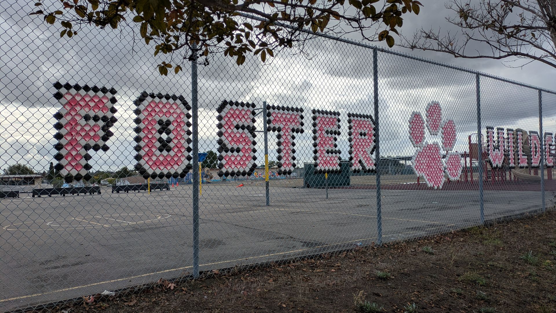 Chain-link fence with pink and black letters spelling "POSTER" and a pink paw print, with a cloudy sky and empty playground behind the fence.