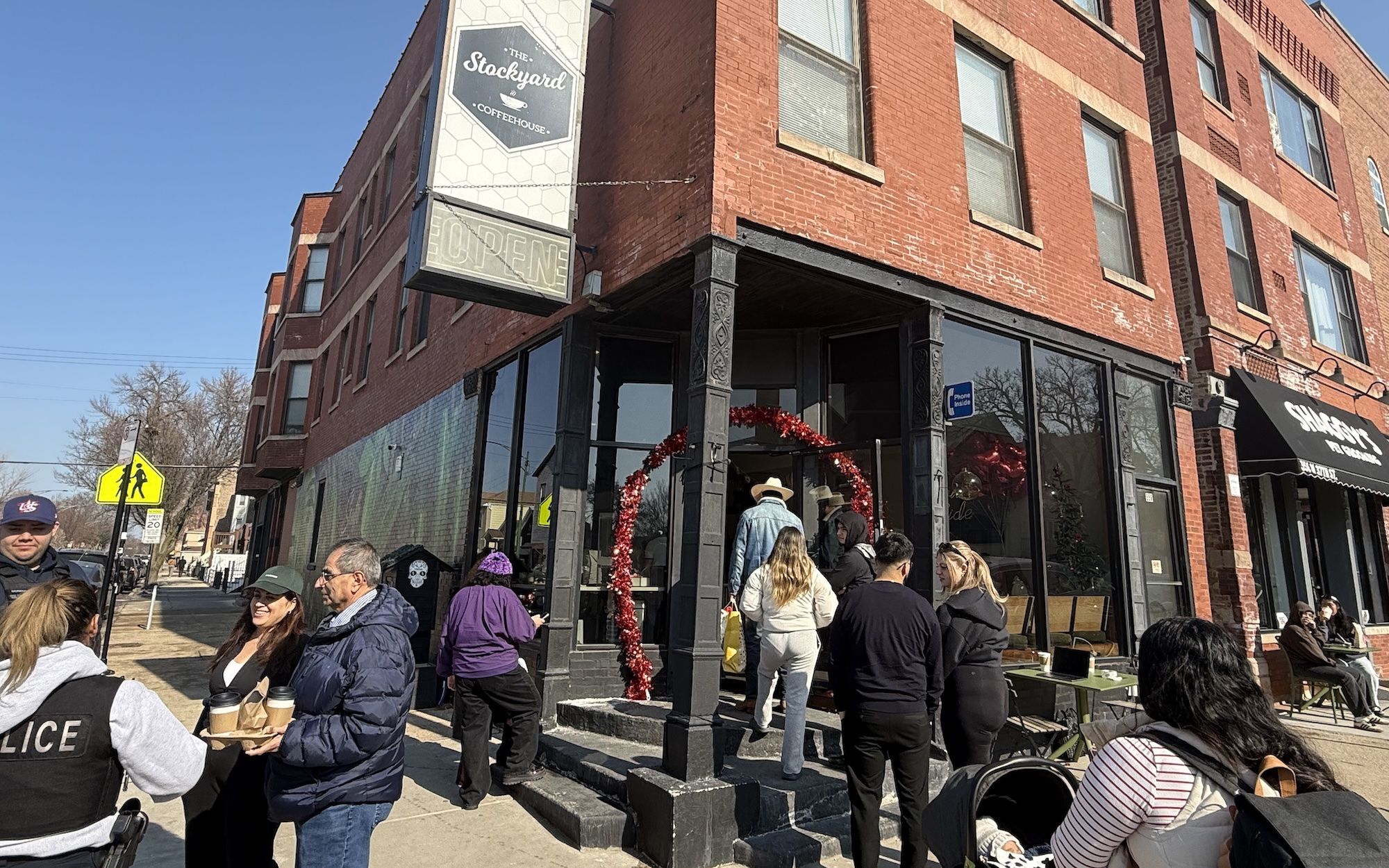 People waiting and entering The Stockyard Coffeehouse, a brick building with a red tinsel arch on the entrance. A police officer talks to customers holding coffee cups outside.