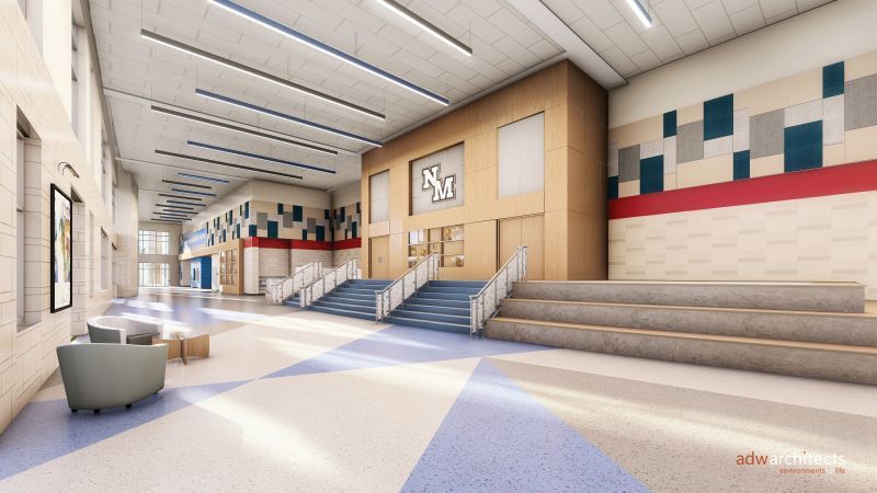Bright, modern school hallway with blue and gray floor patterns, beige walls with red and blue accents, central stairs leading to doors marked "NM", and cozy seating area on the left.