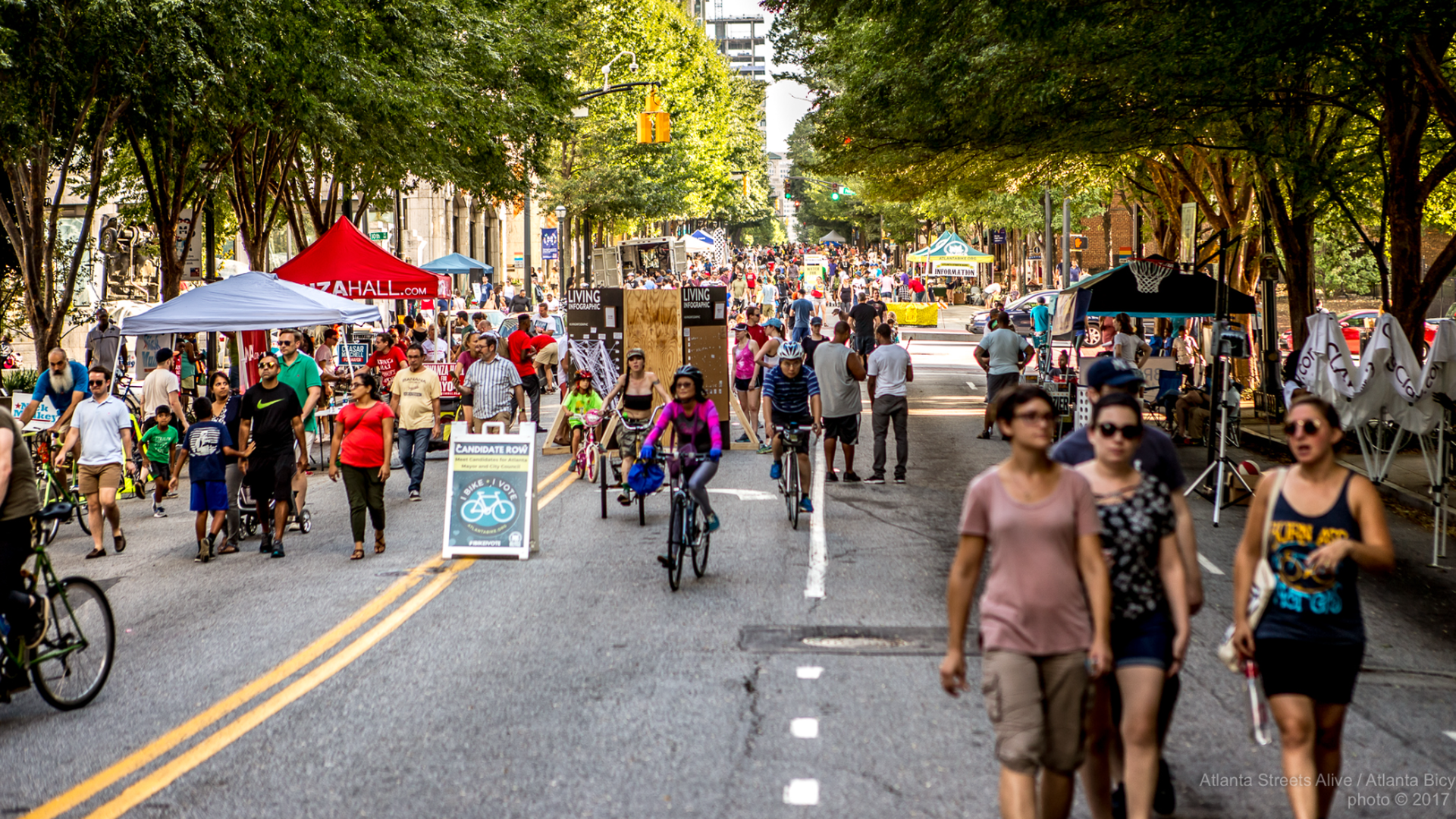 People walk and bike at a street festival shaded by trees and closed to cars in a dense urban setting