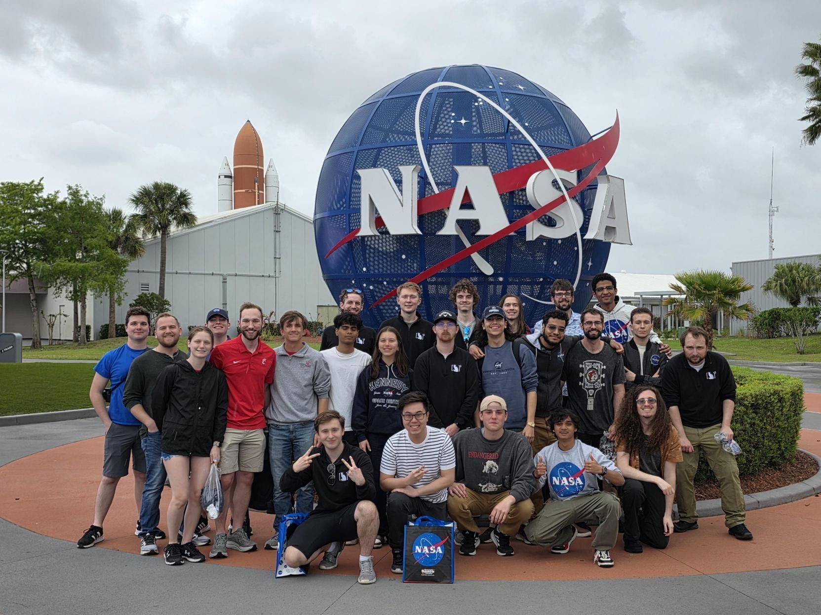 Members of the UC CubeCats team posing in front of a large blue NASA globe sculpture with a red orbital swoosh; rockets and campus buildings in the background.