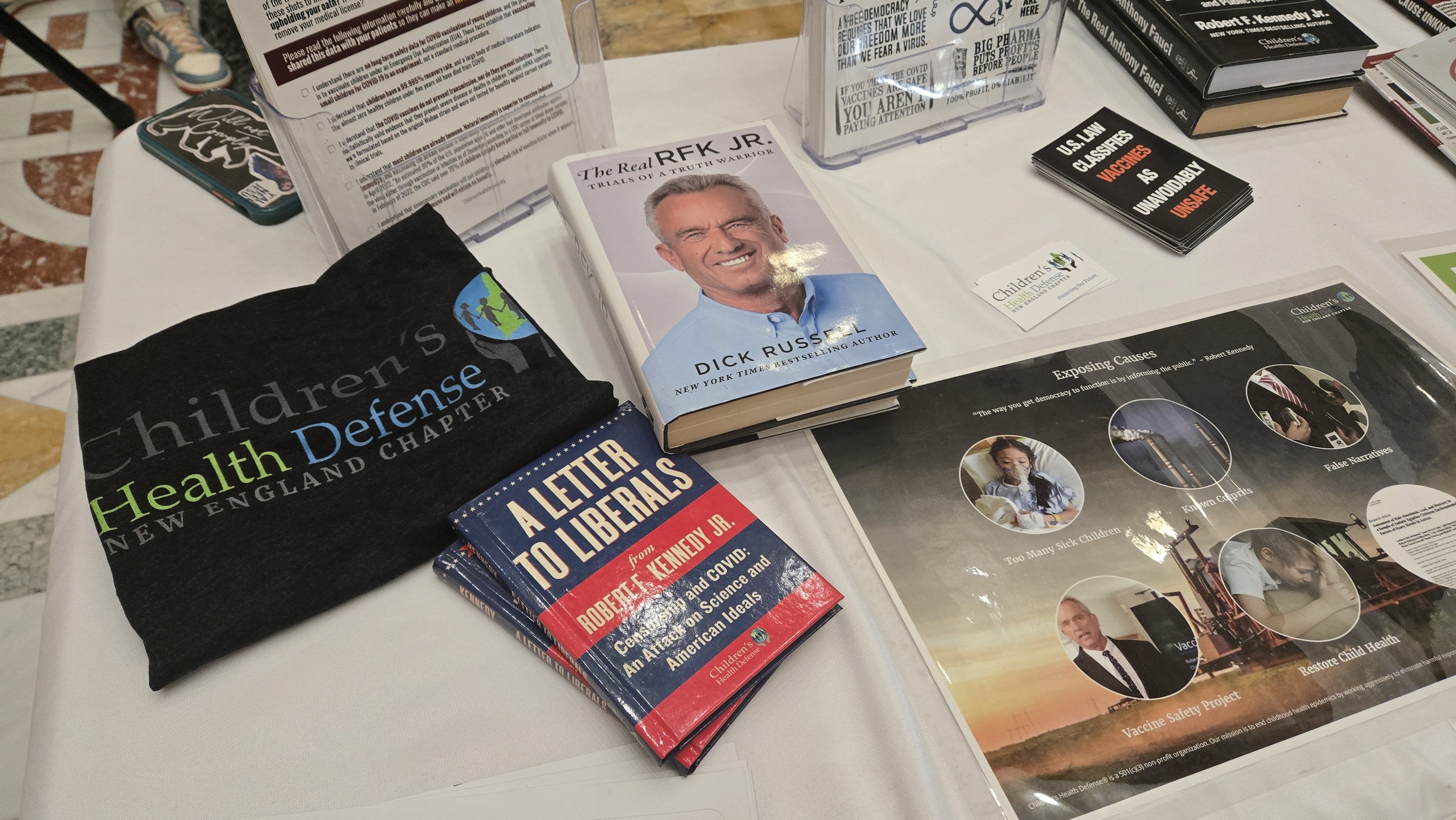 A table displaying books by Robert F. Kennedy Jr. and Dick Russell, a Children's Health Defense shirt, pamphlets, and a poster exposing vaccine safety concerns and related topics.