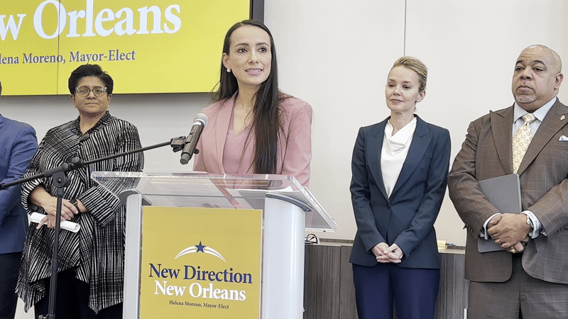 Image shows a woman with long dark hair speaking at a podium. The sign on the podium says "New Direction New Orleans."