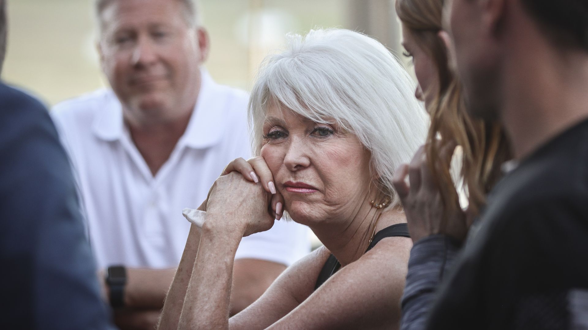 An older woman with short white hair sits outdoors, chin resting on clasped hands, looking contemplative as a small group chats in the background.