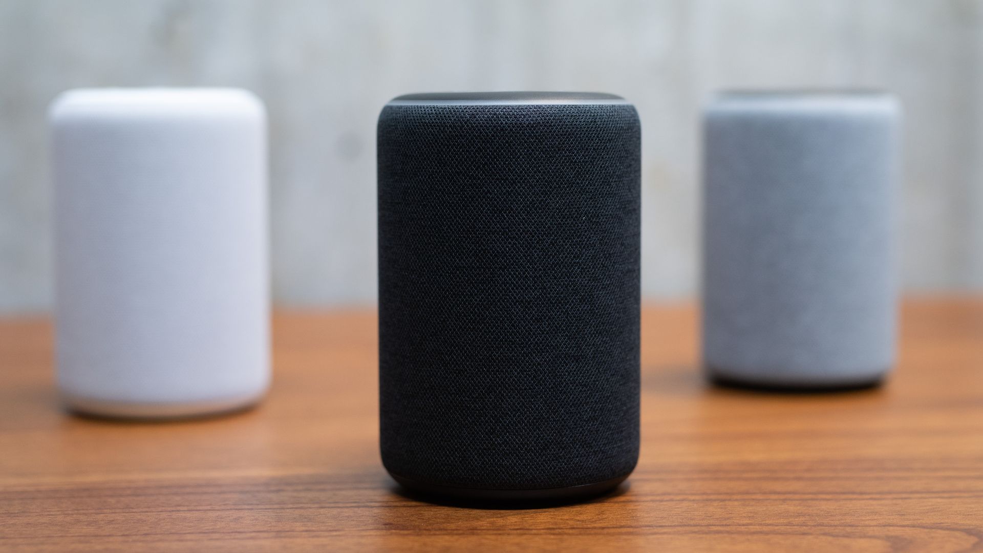 In this image, a white Amazon Alexa sits next to a black one and a gray one on a wooden countertop. 